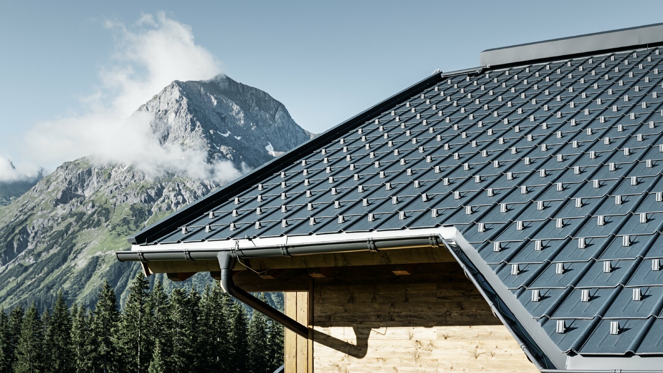 PREFA Dachplatte im alpinen Einsatz - Dachplatte in Anthrazit verlegt auf Hütte in Lech am Arlberg mit Berge im Hintergrund; PREFA Dachrinne in Anthrazit; Holzfassade