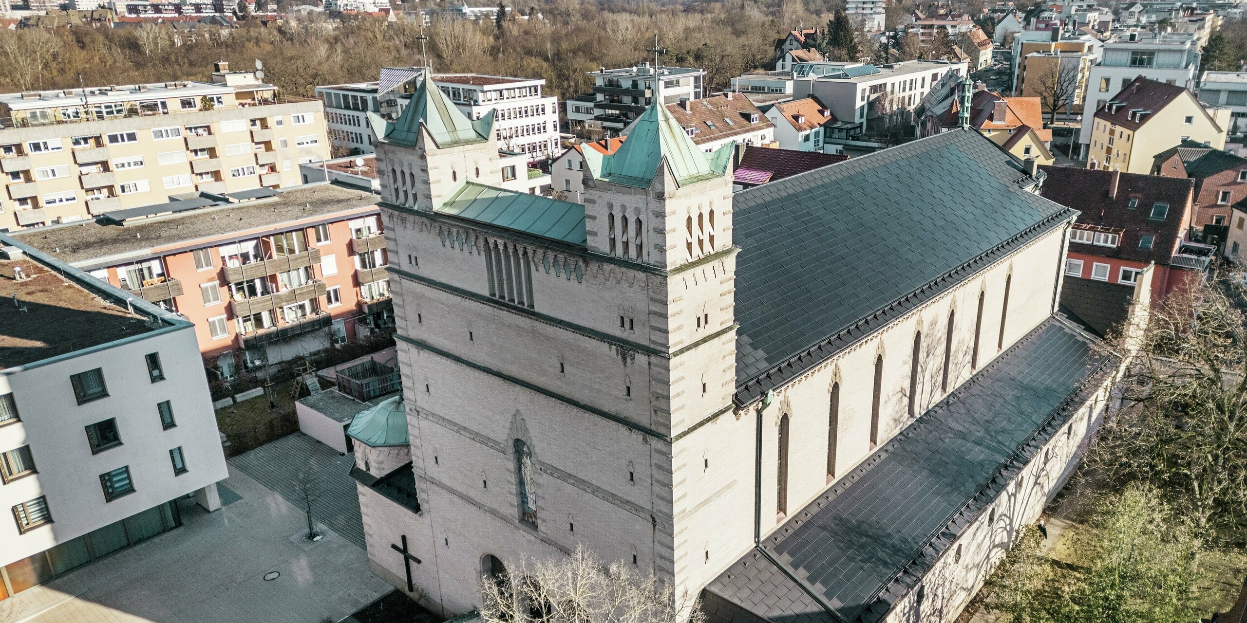 Vorderseite der St. Karl Borromäus Kirche in Nürnberg: Das Bild zeigt das beeindruckende Dach der denkmalgeschützten Kirche, das mit schwarzen PREFA-Solardachplatten gedeckt ist. Die Solarmodule fügen sich harmonisch in die historische Architektur der Kirche ein und stellen gleichzeitig eine moderne Lösung zur nachhaltigen Energiegewinnung dar. Im Hintergrund sind moderne Gebäude und die grünen Hügel Nürnbergs zu sehen, was den gelungenen Übergang zwischen traditioneller Kirchenarchitektur und zeitgemäßer Energieversorgung verdeutlicht. Das Aluminiumdach der Kirche wird durch die stilvolle PREFA Dachplatte R.16 vervollständigt.