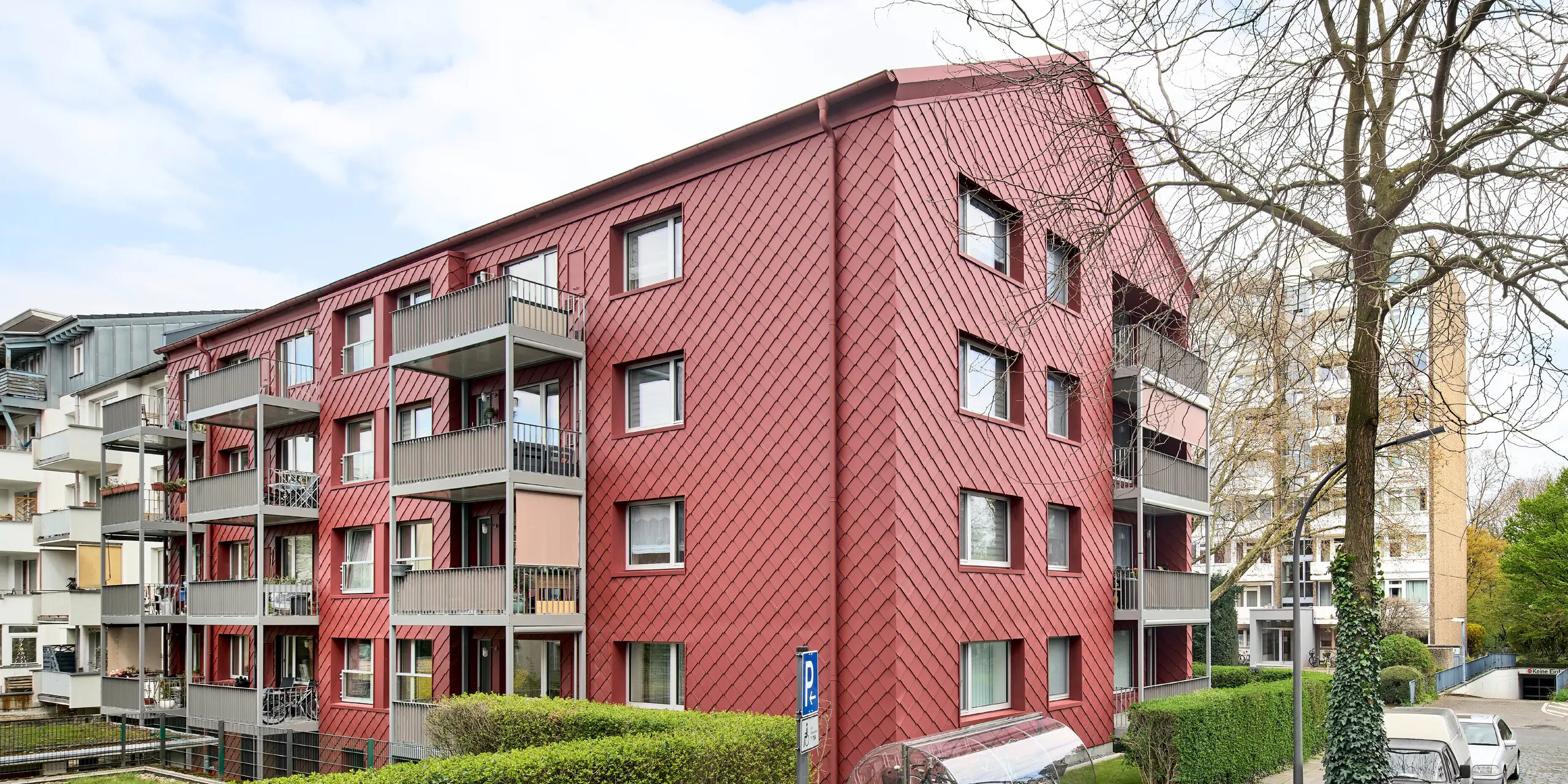 Beeindruckende Gesamtansicht des sanierten Mehrfamilienhauses „Energiesprong 2426“ in Köln-Zollstock: PREFA Wandrauten 29 × 29 in P.10 Oxydrot verleihen der weitläufigen Fassade eine unverwechselbare, lebendige Optik. Das markante Rautenmuster, moderne Fensterreihen und farblich abgestimmte Dachentwässerung setzen architektonische Akzente. Die leichte, langlebige und witterungsbeständige Aluminiumfassade verbindet Nachhaltigkeit mit minimalem Wartungsaufwand.