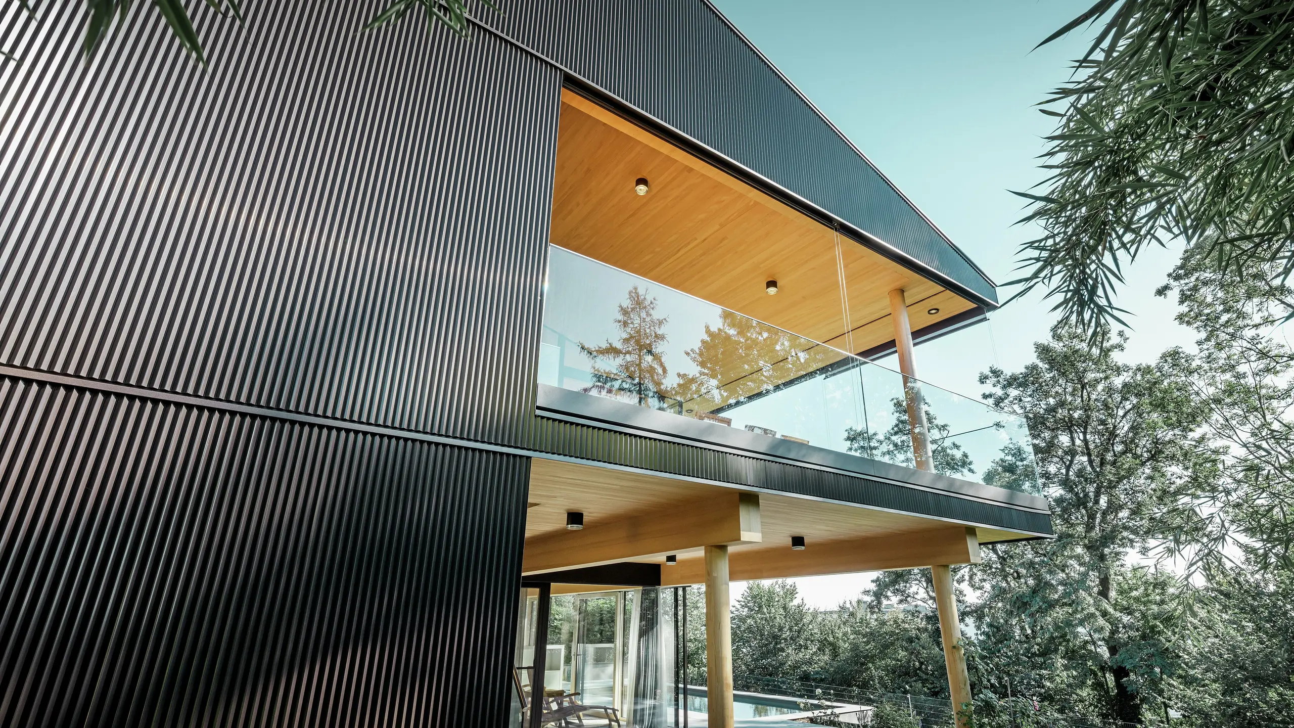 Seitliche Ansicht eines Einfamilienhauses, die Fassade ist mit dunklem PREFA Zackenprofil verkleidet. Man sieht die Terrasse und einen großzügigen Balkon. Die Decke bzw. die Untersicht ist mit Holzpaneelen gestaltet.