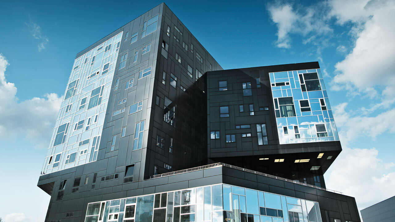 Modernes Universitätsgebäude mit markanter, geometrischer Architektur und überhängenden Baukörpern. Die Fassade besteht aus dunklen und hellen Aluminium-Verbundplatten in unterschiedlichen Grautönen mit unregelmäßig angeordneten Fenstern. Das Gebäude steht unter blauem Himmel mit vereinzelten Wolken und spiegelt das Licht in verschiedenen Facetten wider.