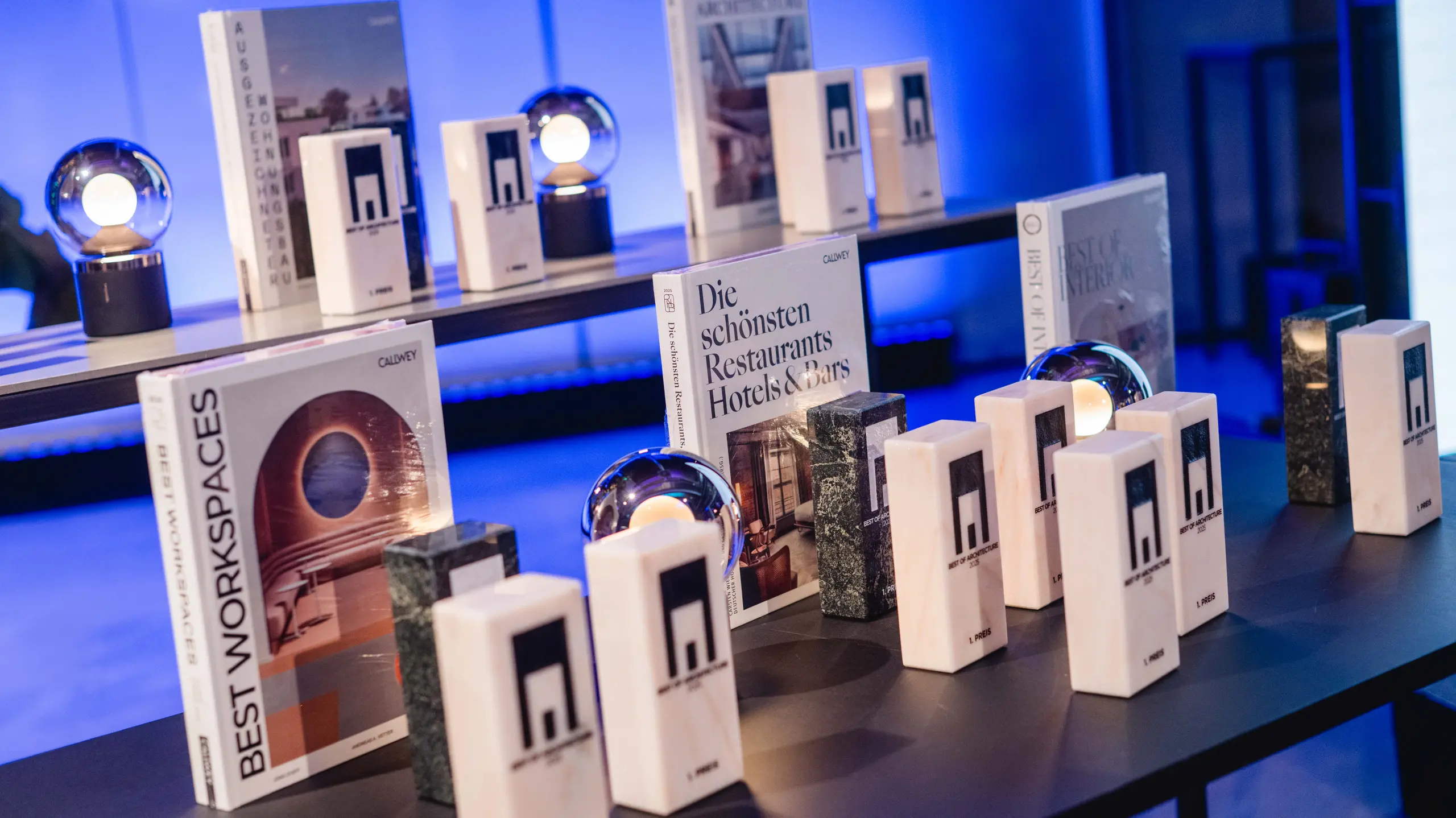 Eine Ausstellung der Auszeichnungen aus dem Wettbewerb „Wohngebäude des Jahres“ 2025: Mehrere elegante Trophäen mit dem Logo „Best of Architecture“ sind auf einem modernen Tisch ausgestellt, neben hochwertigen Büchern über Architektur und Design wie „Best Workspaces“ und „Die schönsten Restaurants Hotels & Bars“. Die Szene ist in stimmungsvolles blaues Licht getaucht, das Exklusivität, Innovation und architektonische Exzellenz vermittelt.