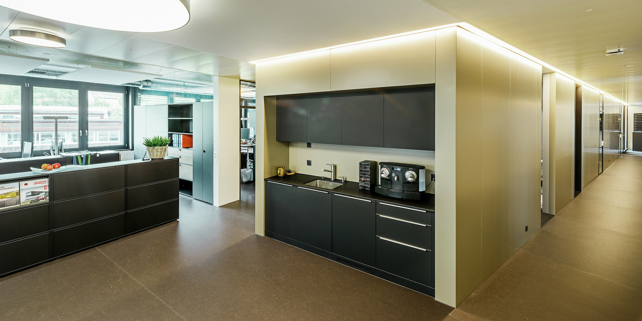 Der Innenbereich des modernen Firmengebäudes in Wabern zeigt einen eleganten Büroflur mit integrierter Kaffeeküche. Die hochwertige Innenverkleidung - bestehend aus der PREFA Aluminium Verbundplatte in Bronze - unterstreicht die klare und zeitlose Architektur. Die indirekte Beleuchtung sorgt für eine angenehme Raumatmosphäre.