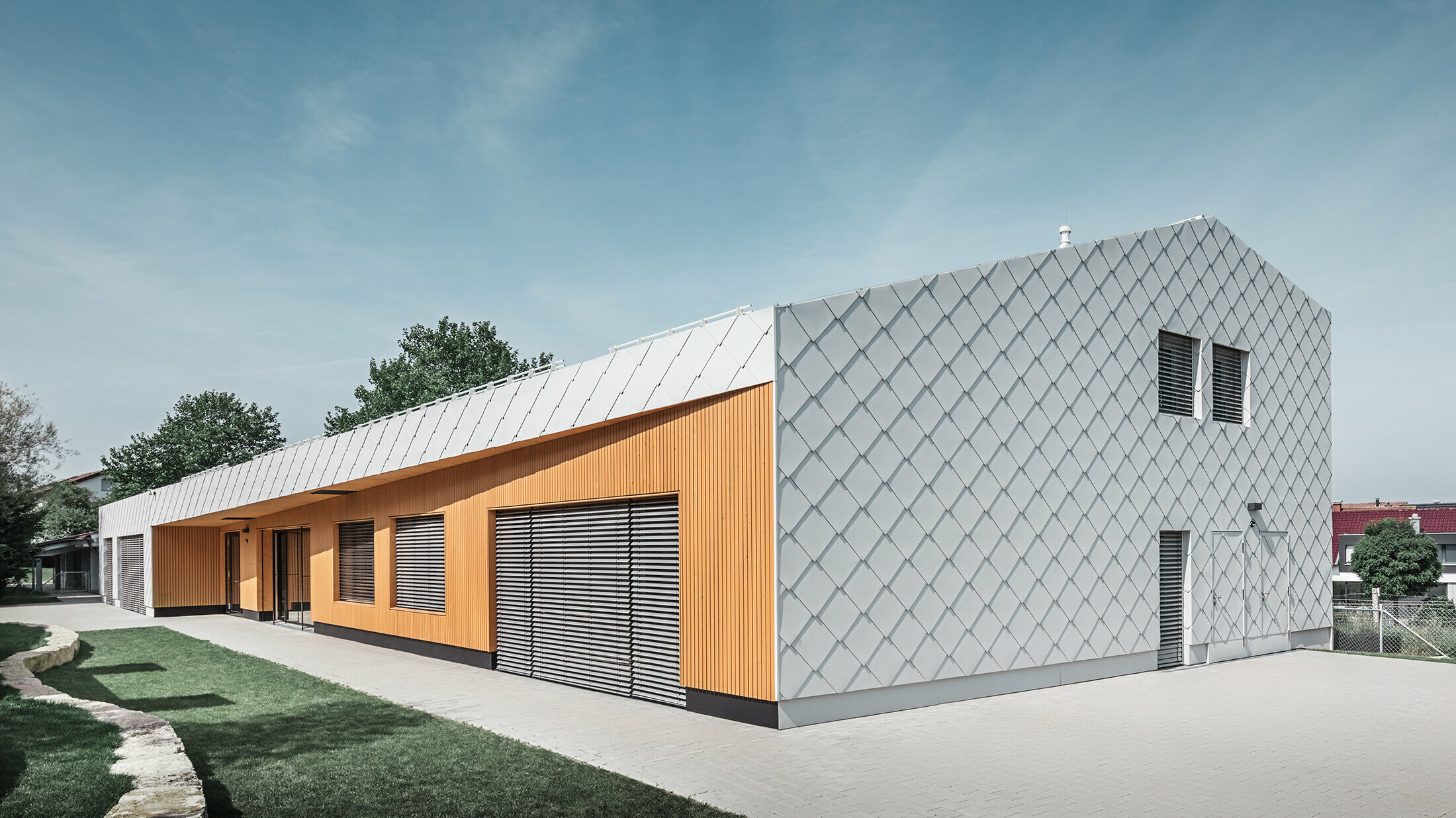 Modernes Kindergartengebäude mit Kombination aus silberner Aluminiumrautenfassade und Holzverkleidung in warmem Orangeton. Die Architektur vereint klare Linien mit funktionalen Elementen wie großen Fenstern und Sonnenschutzlamellen. Das Gebäude steht auf einem gepflasterten Gelände mit angrenzender Rasenfläche unter blauem Himmel.