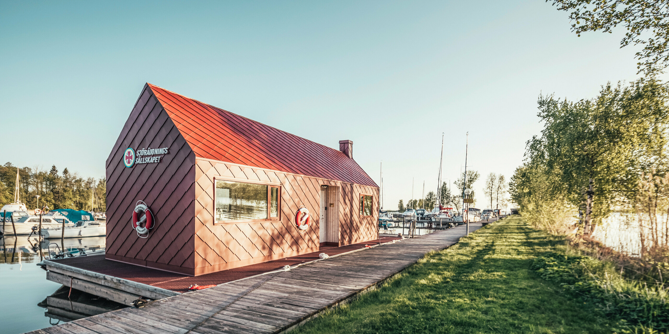 Seenotrettungsstation in harmonischer Umgebung am Hjälmaren See in Schweden, mit einem oxydroten PREFA Dach und Fassadenruten 29×29. Die Station liegt am Ende eines Holzstegs, der in einen Yachthafen führt, wo Boote sanft auf dem Wasser schaukeln. Grüne Bäume rahmen das Bild, das die ruhige, aber bereitwillige Atmosphäre einer Rettungsstation bei goldenem Morgenlicht einfängt.