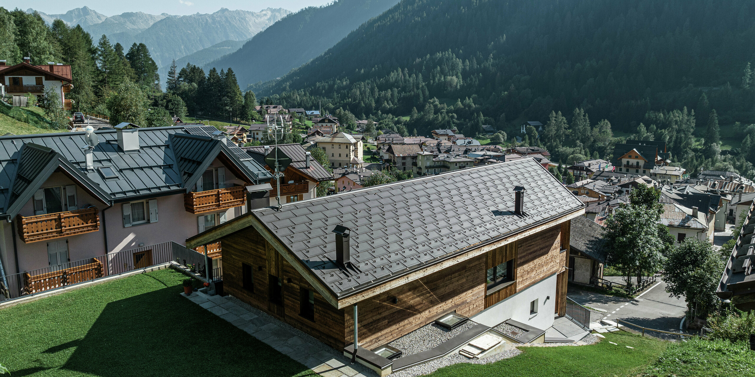 Die Berghütte in Cogolo, Italien, fügt sich mit ihrer Holzfassade und dem PREFA Aluminiumdach in P.10 Braun perfekt in die alpine Landschaft ein. Die widerstandsfähige Dachplatte R.16 schützt zuverlässig vor Schnee, Wind und Regen und sorgt für eine lange Lebensdauer. Eine nachhaltige, stilvolle Lösung für extreme Wetterbedingungen in den Bergen. Im Hintergrund ist das beeindruckende Bergpanorama des Val di Sol zu erkennen.