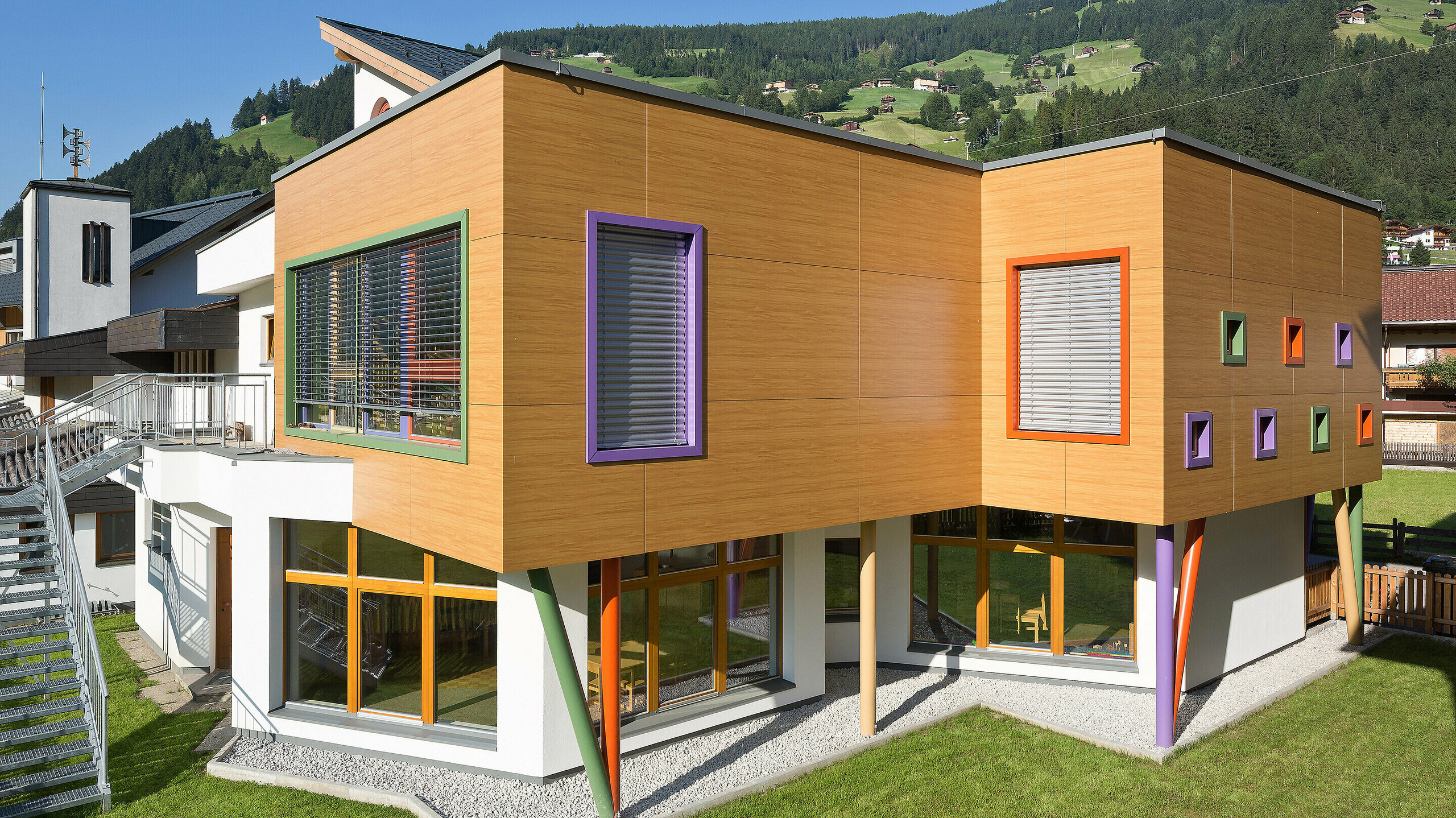 Modernes, zweigeschossiges Gebäude mit auffälliger Holzoptik-Fassade und farbenfrohen Akzenten. Fensterrahmen in Grün, Lila und Orange setzen lebendige Kontraste, während schräge, bunte Stützen die obere Etage tragen. Das Gebäude steht auf einer gepflegten Rasenfläche in einer alpinen Umgebung mit grünen Hügeln und Häusern im Hintergrund.