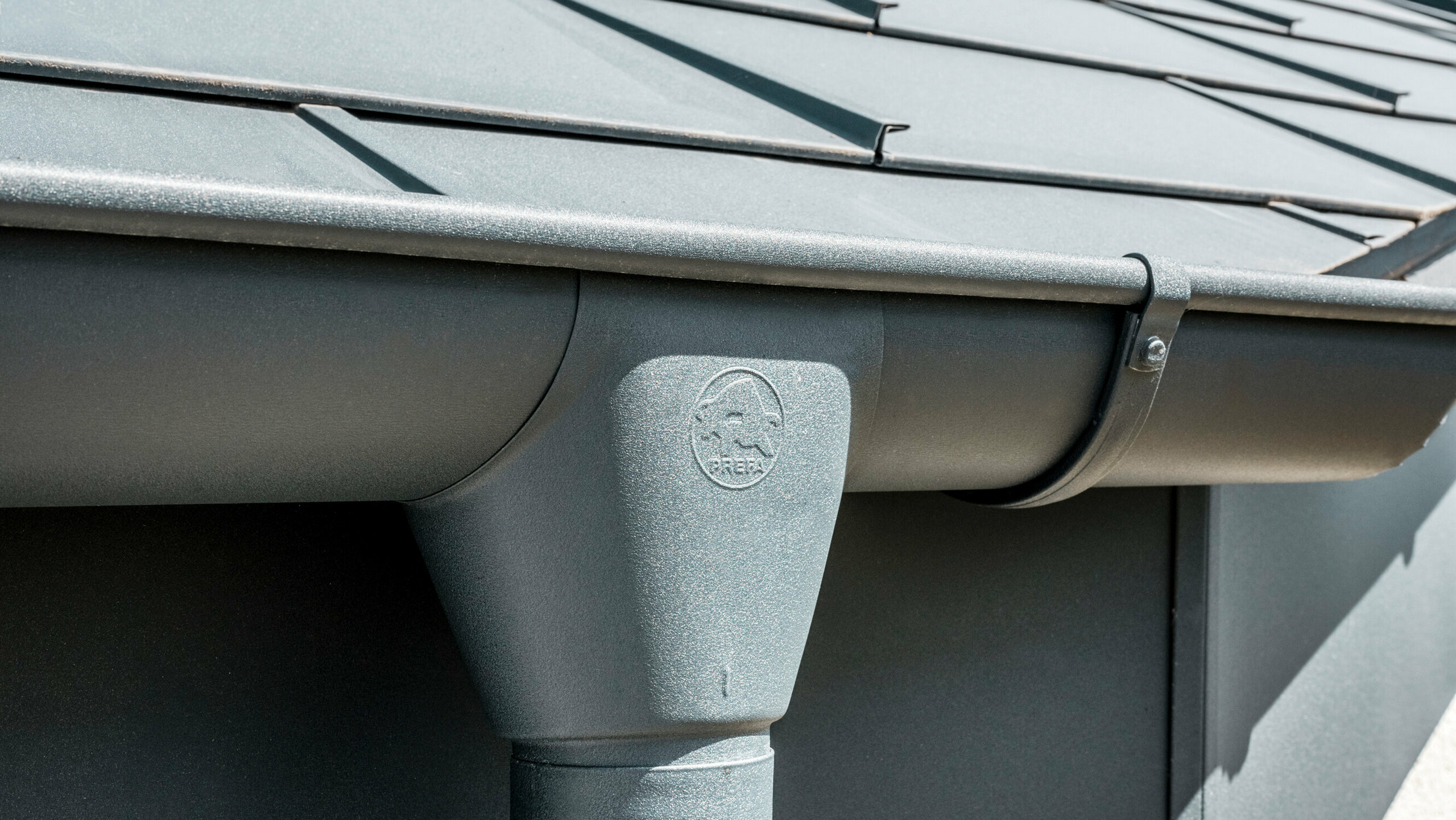 Nahaufnahme eines Hausdaches mit PREFA Dachplatte R.16 in Anthrazit sowie detailreicher Ansicht der farblich abgestimmten PREFA Dachrinne und Fallrohr aus Aluminium bei sonnigem Wetter.