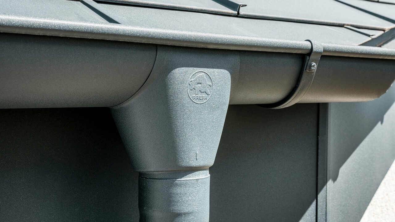 Nahaufnahme eines Hausdaches mit PREFA Dachplatte R.16 in Anthrazit sowie detailreicher Ansicht der farblich abgestimmten PREFA Dachrinne und Fallrohr aus Aluminium bei sonnigem Wetter.