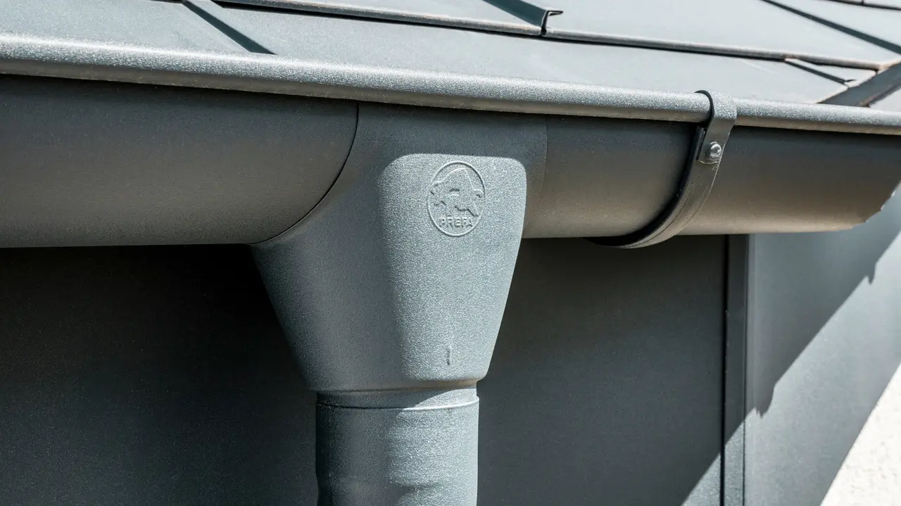 Nahaufnahme eines Hausdaches mit PREFA Dachplatte R.16 in Anthrazit sowie detailreicher Ansicht der farblich abgestimmten PREFA Dachrinne und Fallrohr aus Aluminium bei sonnigem Wetter.