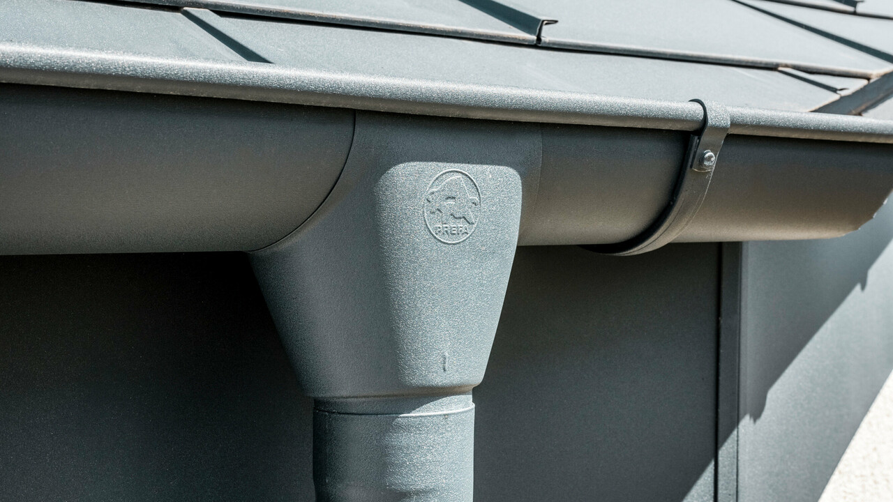 Nahaufnahme eines Hausdaches mit PREFA Dachplatte R.16 in Anthrazit sowie detailreicher Ansicht der farblich abgestimmten PREFA Dachrinne und Fallrohr aus Aluminium bei sonnigem Wetter.