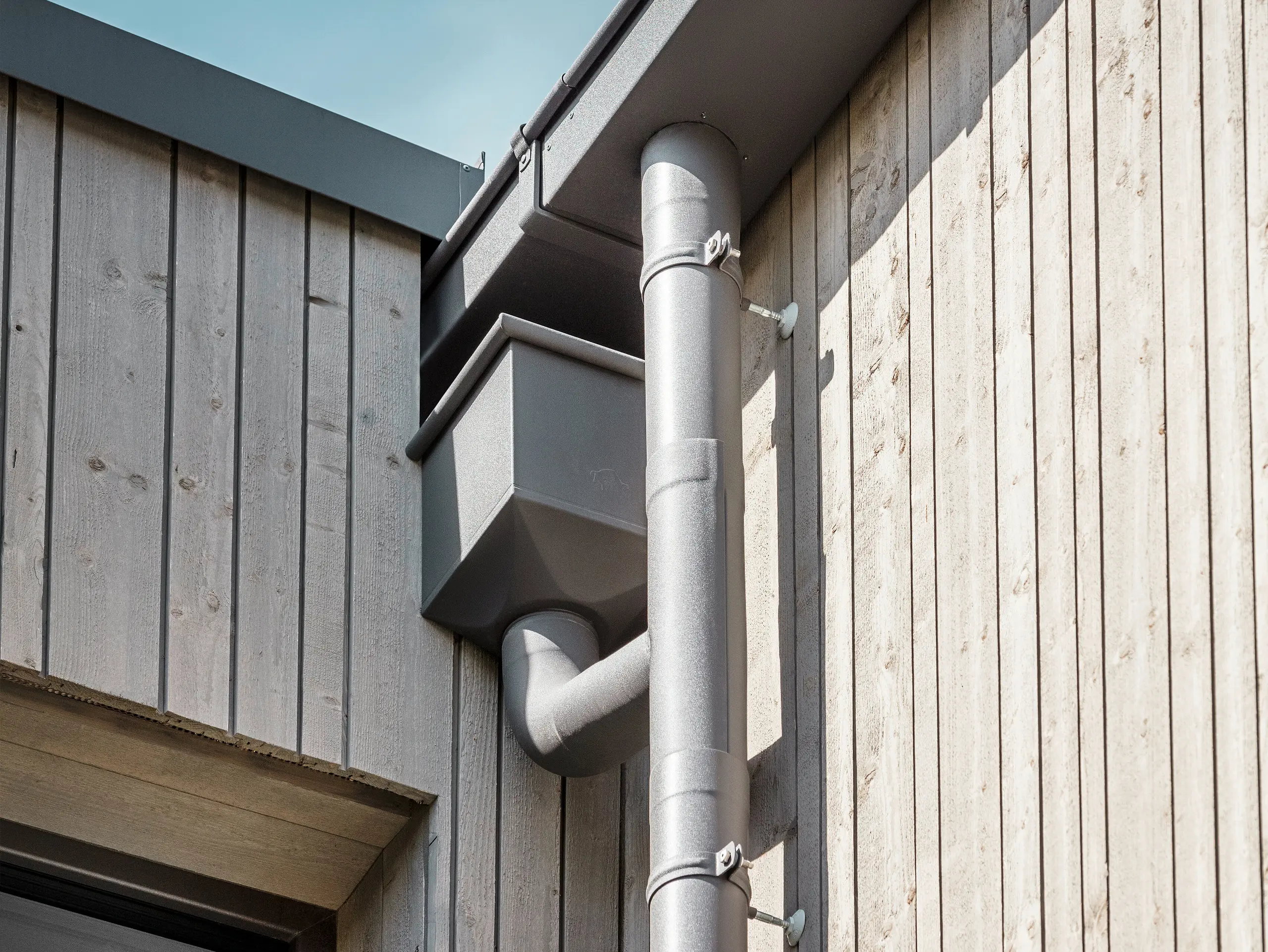 Nahaufnahme einer Holzfassade mit einem PREFA Dachentwässerungssystem aus Aluminium. Der Fokus des Bildes liegt auf dem Ablaufrohr und dem Wasserfangkasten. Das PREFA Dachentwässerungssystem ist in einem hellen Grauton gehalten und schafft einen angenehmen Kontrast zu der natürlichen Holzverkleidung des Gebäudes. 