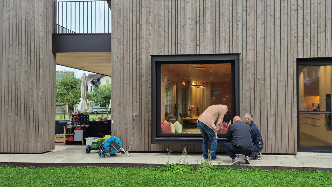 Drei Teilnehmende der PREFA Objekttour begutachten im Detail die Ausführung eines modernen Wohnhauses mit markanter Holzfassade und großzügigen Fensterflächen. Der spannende Kontrast aus natürlichem Holz und präzise verarbeiteten PREFA Aluminium-Fensterbänken und Verblechungen schafft ein harmonisches Gesamtbild. Die langlebigen, wetterbeständigen PREFA Lösungen fügen sich dezent in die zeitgemäße Architektur ein.