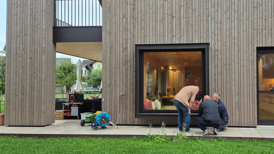 Drei Teilnehmende der PREFA Objekttour begutachten im Detail die Ausführung eines modernen Wohnhauses mit markanter Holzfassade und großzügigen Fensterflächen. Der spannende Kontrast aus natürlichem Holz und präzise verarbeiteten PREFA Aluminium-Fensterbänken und Verblechungen schafft ein harmonisches Gesamtbild. Die langlebigen, wetterbeständigen PREFA Lösungen fügen sich dezent in die zeitgemäße Architektur ein.