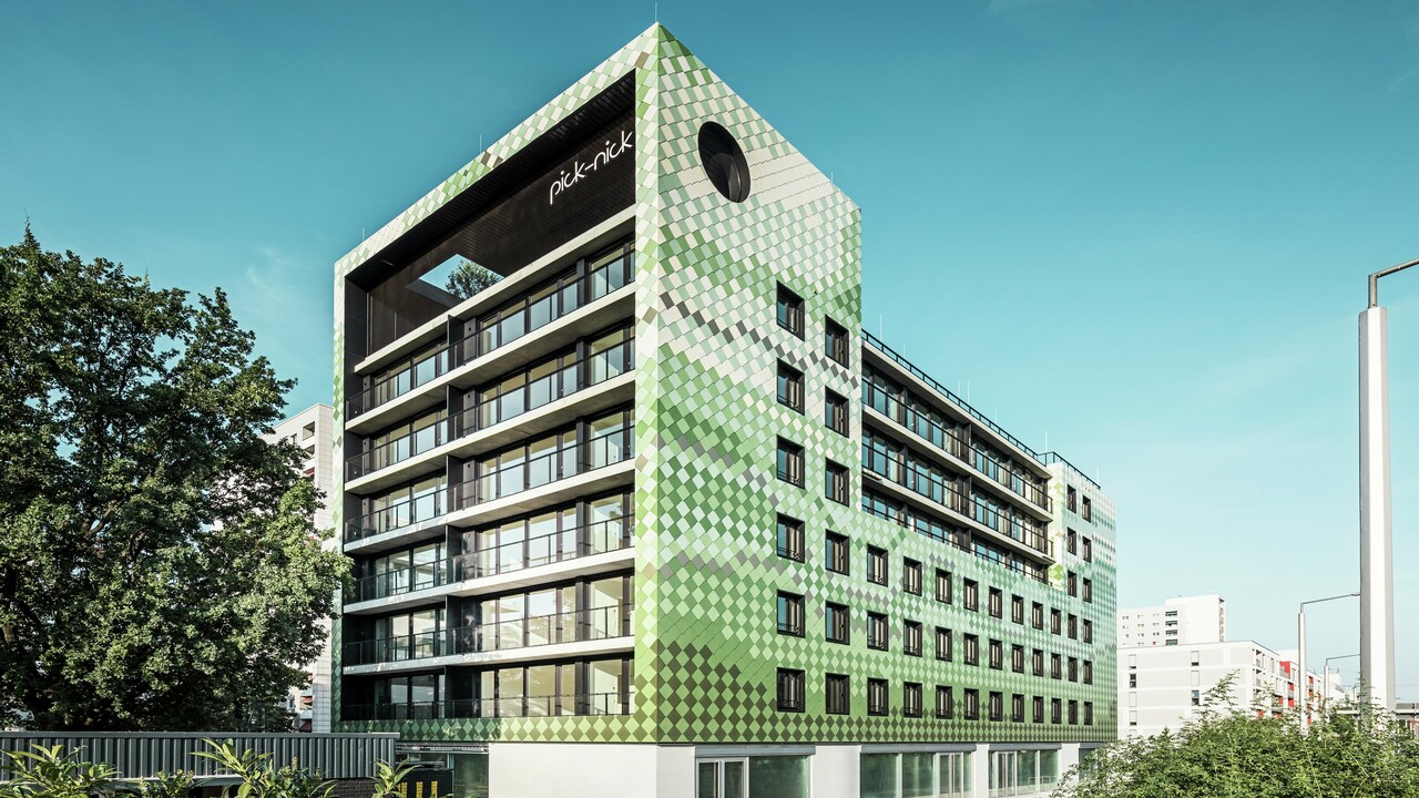 Das Wohn- und Geschäftshaus „Pick-Nick“ in Dresden besticht durch seine moderne PREFA-Aluminiumfassade in verschiedenen Grün- und Beigetönen. Die markante Fassade vereint klare Linien mit einem dynamischen Farbspiel und wird durch große Glasflächen zusätzlich betont. Architektonische Akzente setzen der Schriftzug „Pick-Nick“ an der oberen Gebäudekante und der begrünte Dachgarten.