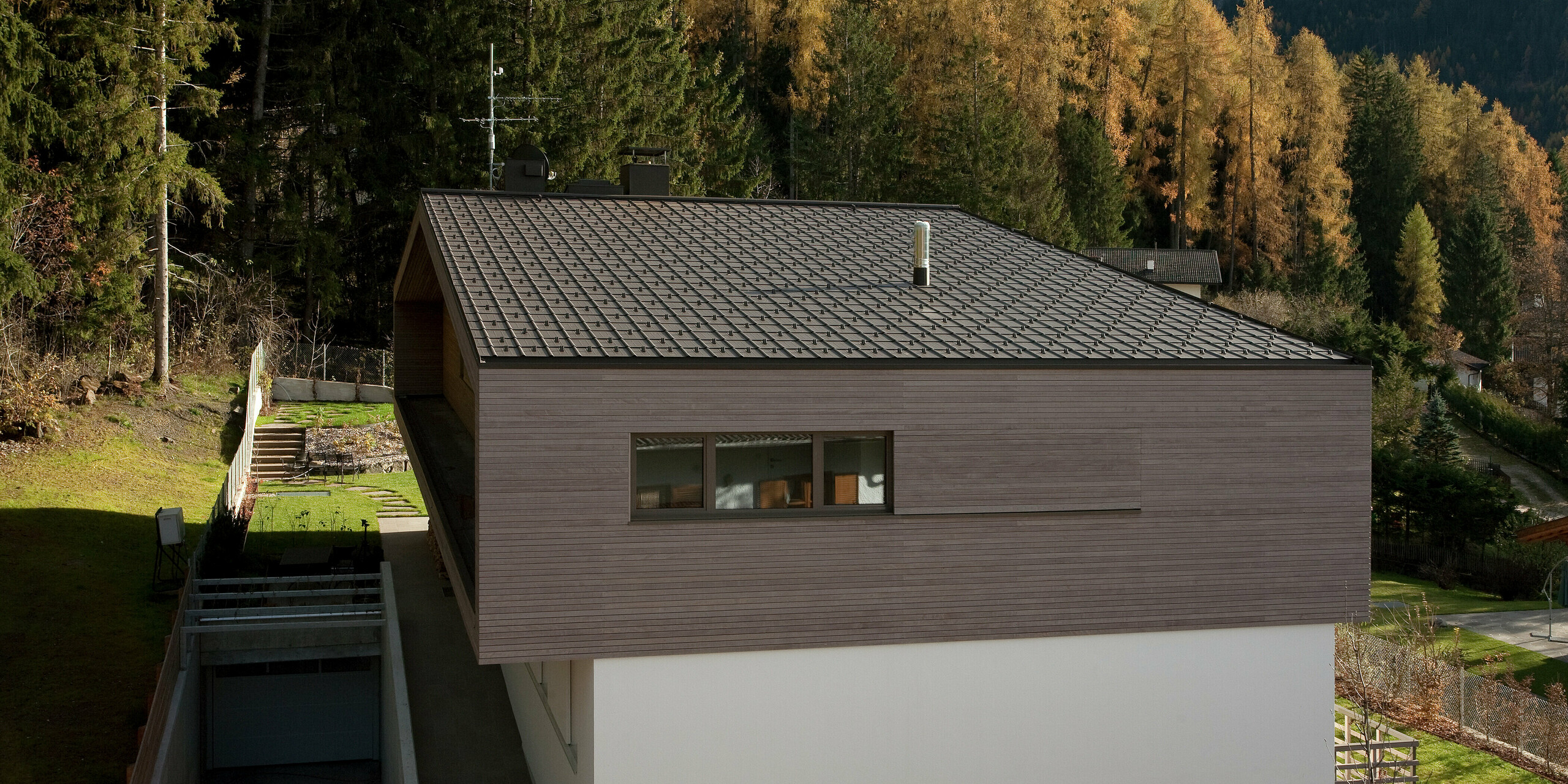 Moderne Architektur mit PREFA Dachplatten in P.10 Braun, umgeben von einem naturnahen Garten und Waldlandschaft. Der minimalistische Baukörper mit Holzfassade fügt sich harmonisch in die Umgebung ein.