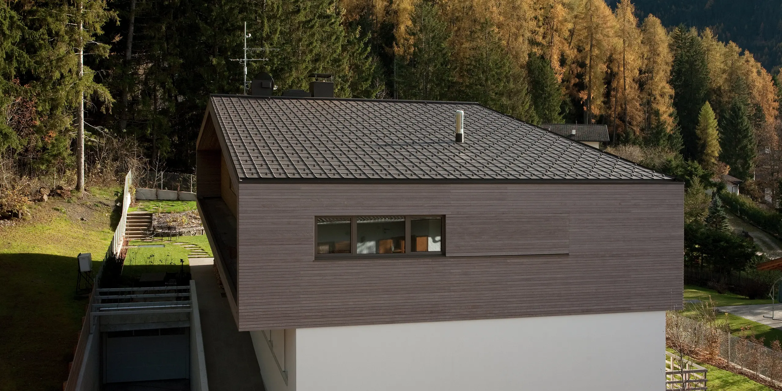 Moderne Architektur mit PREFA Dachplatten in P.10 Braun, umgeben von einem naturnahen Garten und Waldlandschaft. Der minimalistische Baukörper mit Holzfassade fügt sich harmonisch in die Umgebung ein.