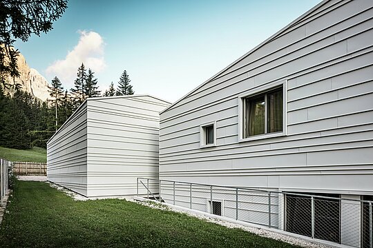 Die Rückseite der Kaserne „Tempesti“ in Corvara bietet eine anschauliche Perspektive: Waldbäume, ein strahlend blauer Himmel und von der Sonne hervorgehobene Berge sind zu sehen. Die helle Aluminiumfassade mit ihrer klaren Struktur zeigt die technische Raffinesse von PREFALZ in der Farbe P.10 Prefaweiß. Die horizontalen Aluminiumscharen reflektieren das Licht, bieten optimalen Witterungsschutz und fügen sich harmonisch in die alpine Umgebung ein. Ein Paradebeispiel für langlebige und moderne Militärarchitektur!