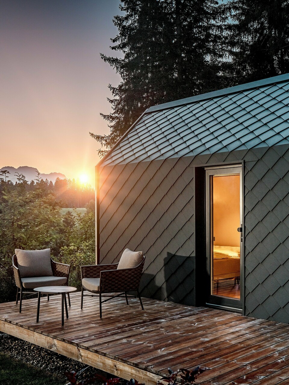 Ein minimalistisches Tiny House im Secret Koi Garden am Natterer See zeigt sich bei Sonnenuntergang mit vorgelagerter Holzterrasse und Blick in den Wald. Dach und Fassade sind mit kleinteiligen PREFA Dach‑ und Wandrauten aus Aluminium in P.10 Anthrazit ausgeführt, deren strukturierte Oberfläche das warme Licht eindrucksvoll reflektiert. Das langlebige, witterungsbeständige PREFA Aluminium schützt die Architektur zuverlässig und schafft einen markanten Kontrast zu Holz, Natur und sanfter Beleuchtung.