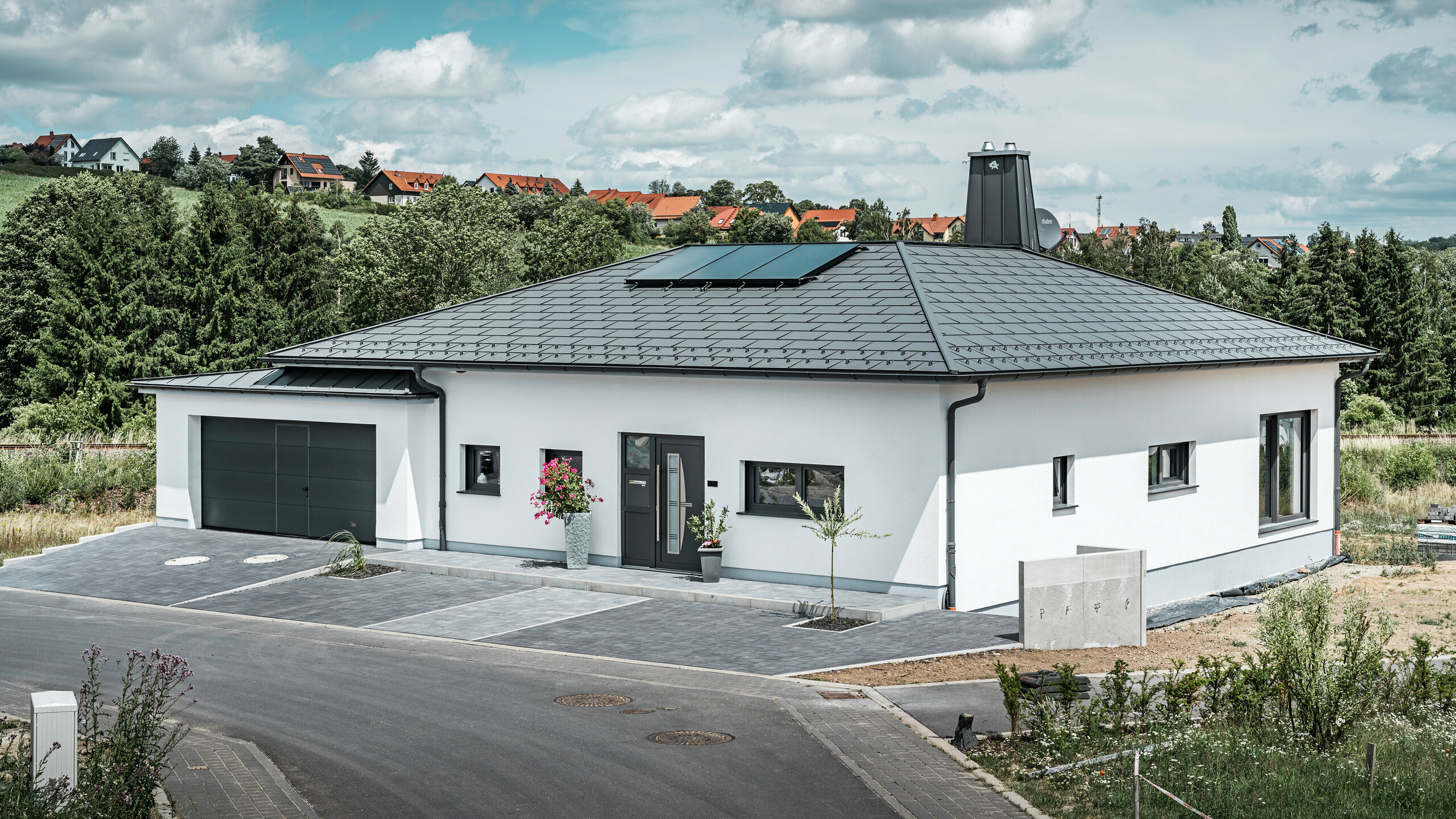 Elegantes Einfamilienhaus in Ilmenau, Thüringen, mit einem hochwertigen PREFA Dach in der Farbe P.10 Dunkelgrau, ausgestattet mit der robusten Dachplatte R.16. Die Garage harmoniert in Farbe und Stil durch die Verwendung von PREFALZ. Der Blickfang des Anwesens ist das funktional gestaltete Atrium und der mit dem PREFA Stier verzierte Schornstein, welcher das moderne Design und die innovative Dachtechnologie von PREFA unterstreichen. Die Solarpaneele fügen sich nahtlos in das nachhaltige Konzept des Hauses ein. Abgerundet wird das durchdachte Gesamtkonzept des Bungalows durch das PREFA Dachentwässerungssytem, welches ebenfalls in Dunkelgrau gehalten ist.
