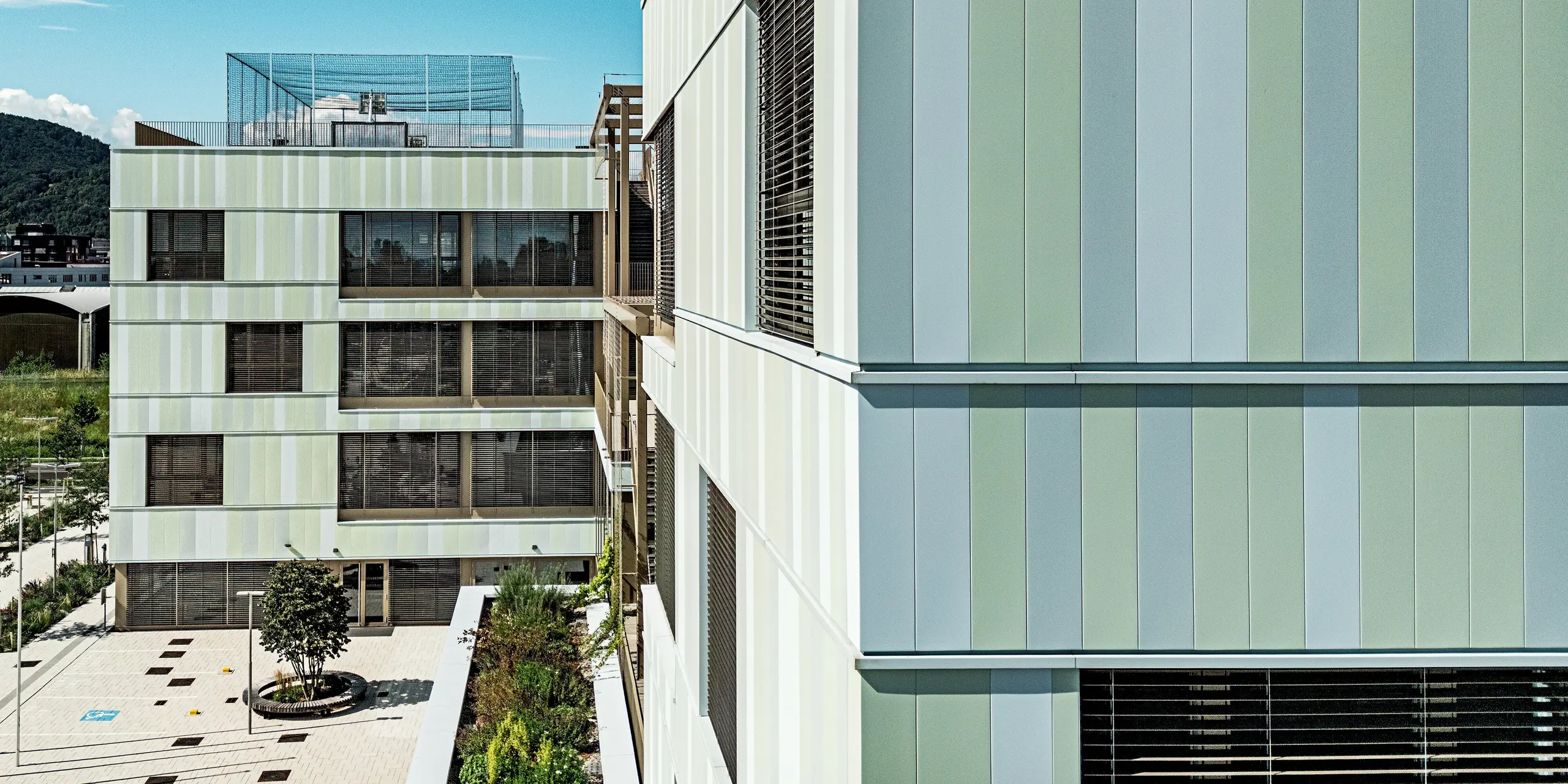 Das Bild zeigt die moderne Fassade der AHS Reininghaus in Graz, die durch ihre auffälligen PREFA-Sidings in Sonderfarben besticht. Die in zarten Grüntönen und Weiß gehaltenen Fassadenelemente verleihen dem Gebäude eine elegante, zugleich aber auch dynamische Ausstrahlung. Die Fenster sind mit dunklen Jalousien ausgestattet, die nicht nur einen funktionalen Sonnenschutz bieten, sondern auch einen ästhetischen Kontrast zur helleren Fassade bilden. Das Gebäude fügt sich perfekt in die umgebende Landschaft ein. Die Kombination aus modernen Materialien und durchdachtem Design macht die Schule zu einem Blickfang in der Grazer Stadtlandschaft.