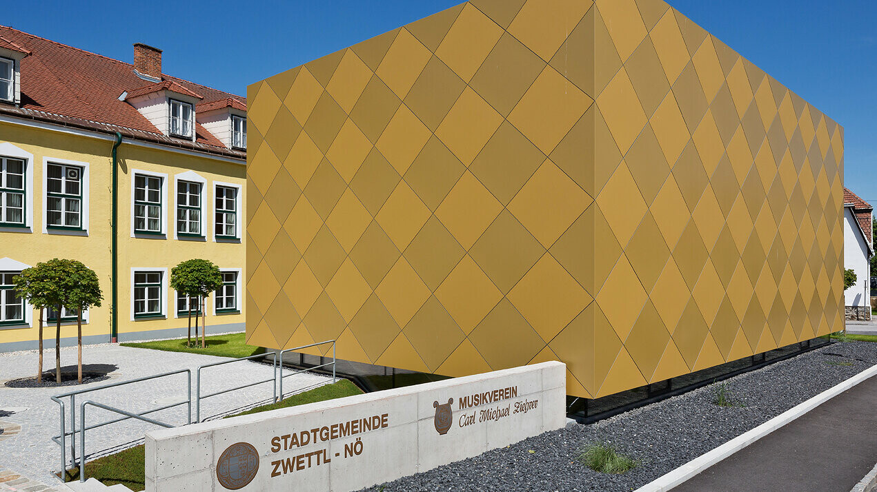 Eingangsbereich zum Musikverein Zwettl. Vor dem Gebäude ist die Funktion des Objekts in Stein gemeißelt. Das quaderförmige Gebäude besticht durch die markante Optik mit PREFA Aluminium Verbundplatten in zwei unterschiedlichen Goldtönen. Die Aluminiumplatten sind sorgfältig in Form von Karos auf der Fassade montiert worden. 