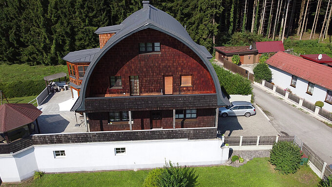 Einfamilienhaus mit markantem Tonnendach, gedeckt mit PREFA Dachschindeln in P.10 Dunkelgrau nach einer Dachsanierung. Das Haus hat eine Fassade aus Holzschindeln und befindet sich in einer ruhigen, grünen Umgebung mit einem angrenzenden Garten und einer Terrasse. Im Hintergrund sind ein Wald und benachbarte Gebäude zu sehen, die an eine schmale Straße angrenzen.