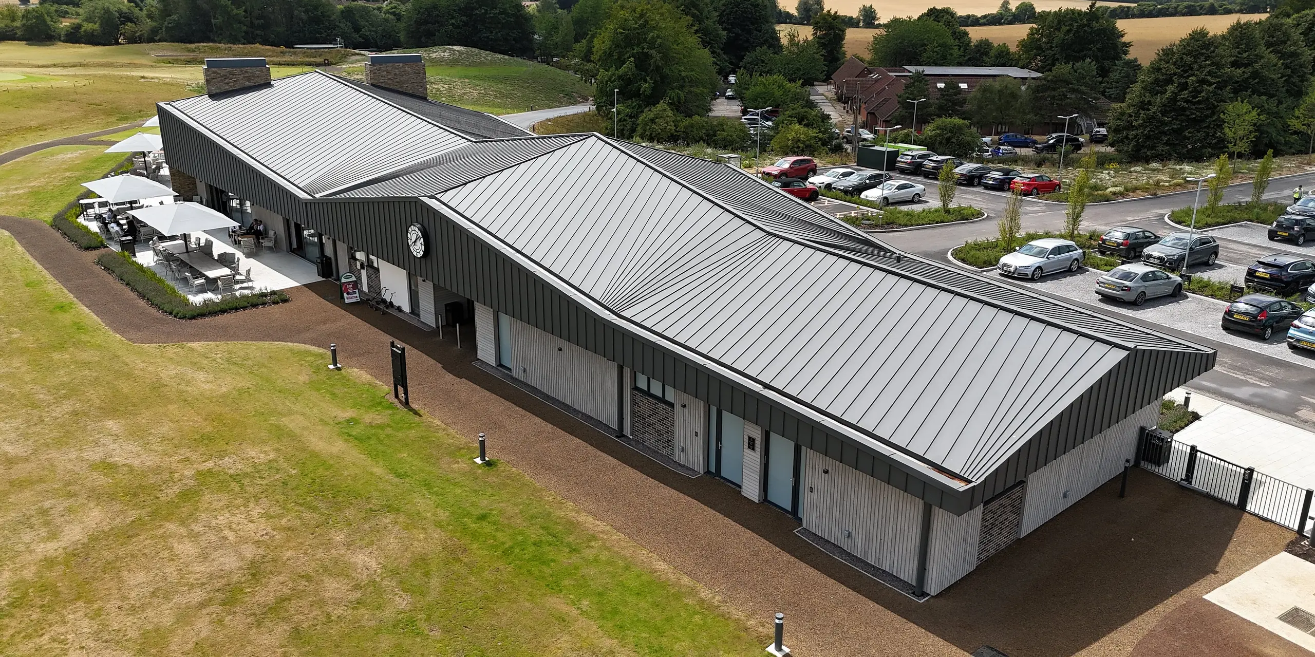 Der Basingstoke Golf Club in Hampshire beeindruckt mit einem modernen Neubau, bei dem rund 1.600 m² PREFALZ Stehfalzbahnen in P.10 Dunkelgrau das Dach und die obere Fassade prägen. Die klaren Linien und die elegante Dachform setzen markante Akzente und harmonieren mit der grünen Umgebung des Golfplatzes. Die untere Fassade aus Holzpaneelen und Ziegeln ergänzt das stilvolle Design.
