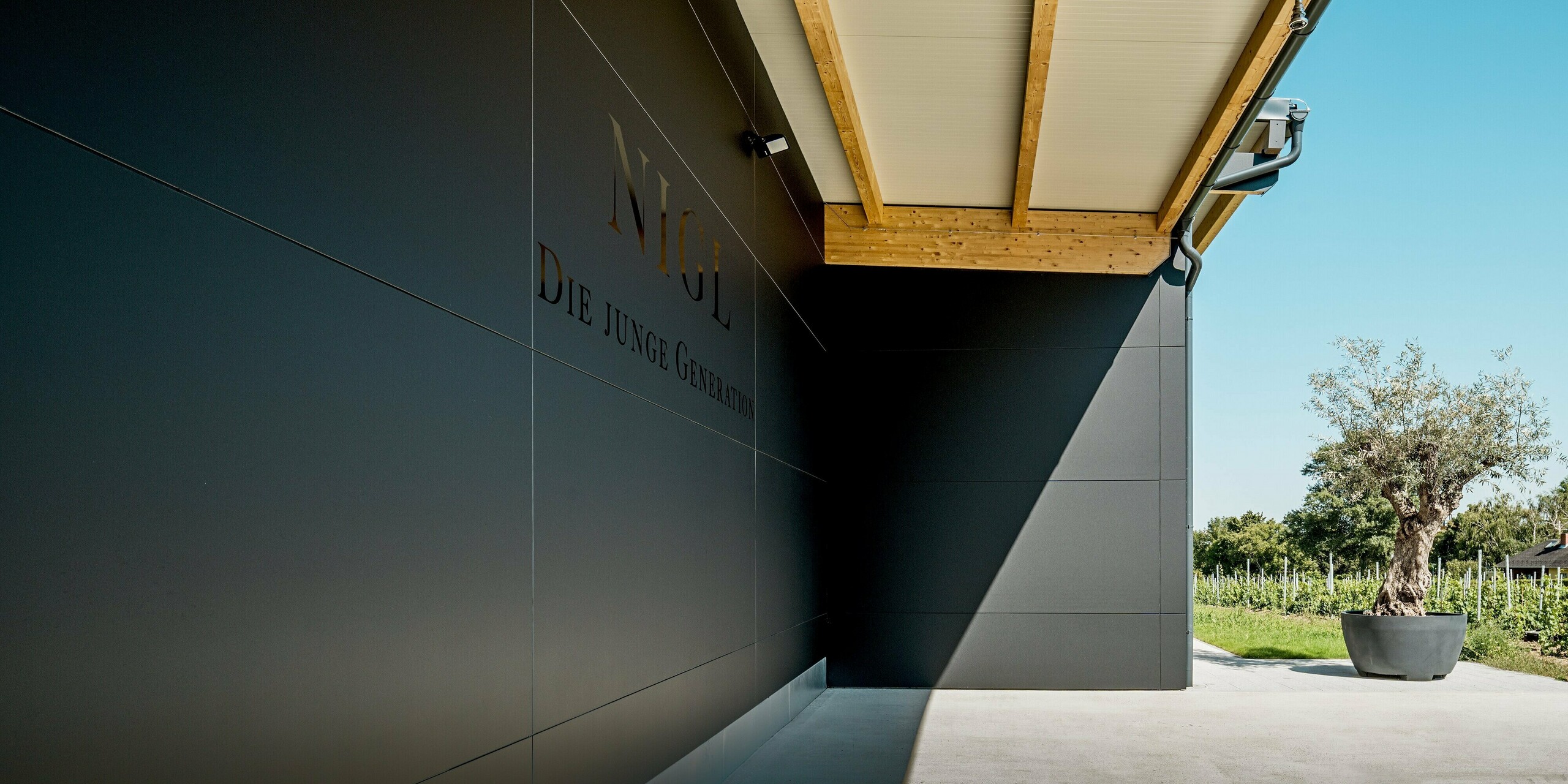 Der Zulieferungsbereich des Weinguts Nigl verfügt über eine markante PREFA Aluminiumfassade in der Farbe P.10 Schwarz. Die glatte, matte Oberfläche der Verbundplatten wirkt elegant und modern, während die exakt ausgeführten Fugenlinien ein klares Fassadenraster bilden. Über dem geschützten Bereich, auf dem der Schriftzug „Nigl – Die junge Generation“ zu lesen ist, spannt sich ein ausdrucksstarkes Vordach mit sichtbarer Holzkonstruktion. Die Kombination aus Aluminium, Holz und Beton erzeugt einen hochwertigen, architektonisch spannenden Kontrast. Rechts im Bild steht ein Olivenbaum im Pflanzgefäß vor der Weinlandschaft – ein Symbol für Naturverbundenheit und Stil. PREFA überzeugt hier durch Design, Dauerhaftigkeit und Wartungsfreiheit.