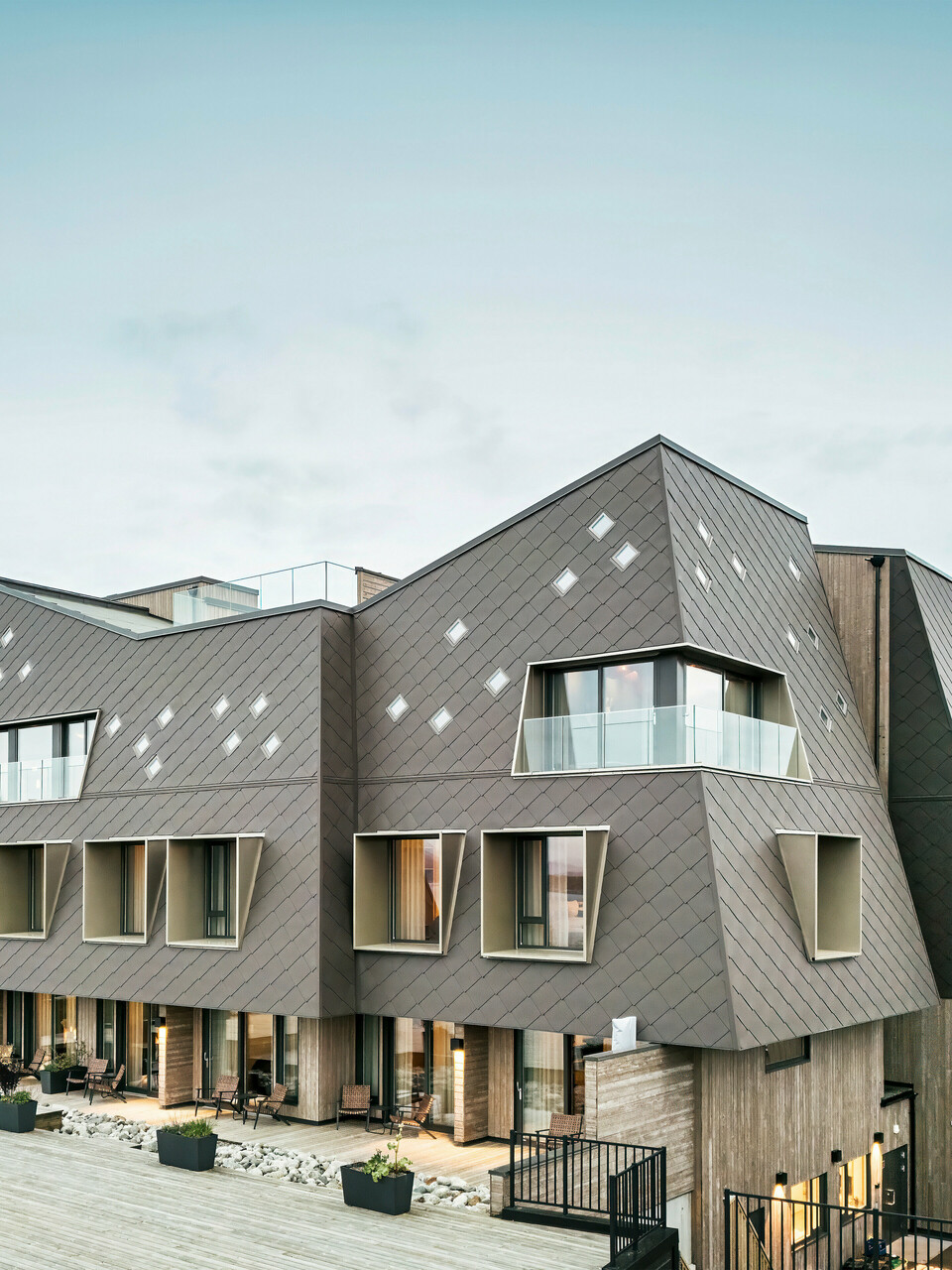 Blick auf die Terrasse des Hotels Beckerwyc House in Bekkjarvik, Norwegen. Das moderne Gebäude wird von einer einer markanten Aluminiumfassade aus PREFA Wandrauten 44 × 44 in Nussbraun geschmückt. Die futuristische Architektur mit schrägen Dachflächen und hervortretenden Fensterelementen wird durch die großzügigen Außenbereiche ergänzt. Die natürliche Farbgebung der Fassade passt sich harmonisch in die umgebende Landschaft ein und unterstreicht das zeitgemäße Design des Gebäudes.