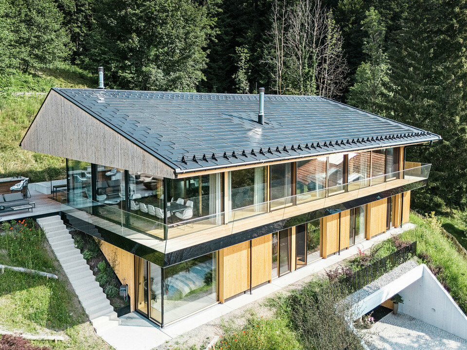 Vogelperspektive auf ein modernes Einfamilienhaus in Egg, Vorarlberg, mit PREFA Solardachplatten und Dachplatten R.16 in P.10 Schwarz. Die Dachkonstruktion aus Aluminium sorgt für nachhaltige Energiegewinnung und bietet robusten Schutz vor Witterungseinflüssen. Der Schneeschutz verhindert in den Wintermonaten abrutschende Dachlawinen. Die großzügige Glasfassade und die Holzverkleidung runden das formschöne Design ab, während im Hintergrund die Terrasse mit hochwertigen Gartenmöbeln zu sehen ist.