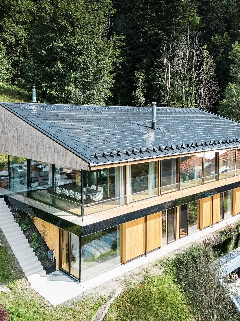 Vogelperspektive auf ein modernes Einfamilienhaus in Egg, Vorarlberg, mit PREFA Solardachplatten und Dachplatten R.16 in P.10 Schwarz. Die Dachkonstruktion aus Aluminium sorgt für nachhaltige Energiegewinnung und bietet robusten Schutz vor Witterungseinflüssen. Der Schneeschutz verhindert in den Wintermonaten abrutschende Dachlawinen. Die großzügige Glasfassade und die Holzverkleidung runden das formschöne Design ab, während im Hintergrund die Terrasse mit hochwertigen Gartenmöbeln zu sehen ist.