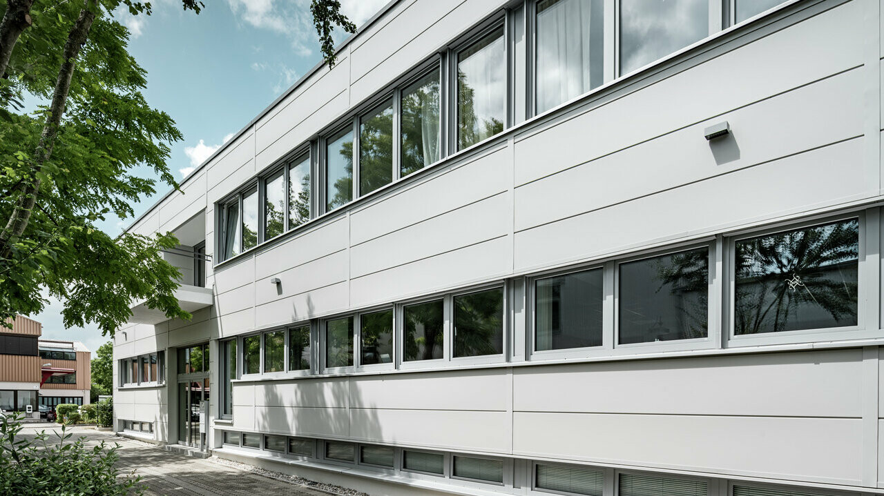 Seitenansicht der PREFA Sidings in der Farbe Prefaweiss, das Objekt hat mehrere Fenster, wobei die Wolken des Himmels gespiegelt werden.
