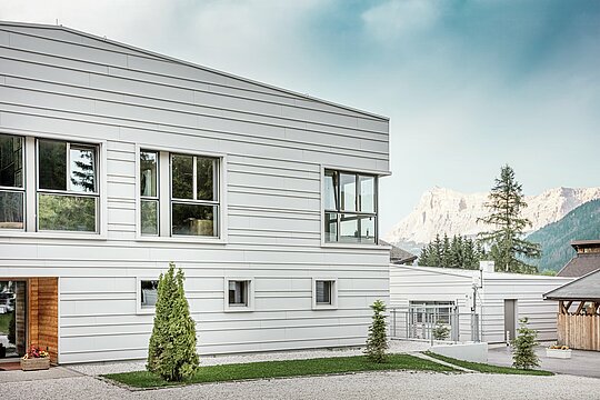 Detailansicht der Kaserne „Tempesti“ mit präzise verlegten PREFALZ-Bahnen in der Farbe P.10 Prefaweiß. Die klaren Linien der horizontal verlegten Stehfalz-Deckung betont die moderne Fassadengestaltung. Die helle Oberfläche reflektiert das Sonnenlicht und optimiert das Mikroklima. Im Hintergrund erhebt sich die beeindruckende Südtiroler Bergwelt – hier trifft Technik auf Natur auf höchstem Niveau.