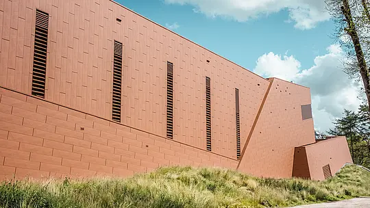 Markante Seitenansicht des Trinkwasserpumpwerks WML in Lottum mit klar gegliedertem, langgestrecktem Baukörper inmitten einer naturnahen Wiesenlandschaft. Die PREFA Siding.X Aluminiumfassade in warmer Rostoptik wird durch rhythmisch gesetzte, schlanke Lamellenöffnungen strukturiert und erzeugt ein lebendiges Spiel aus Licht und Schatten. Die fein facettierte Oberfläche wirkt je nach Perspektive dynamisch und hochwertig. Die leichten, langlebigen und vollständig recycelbaren PREFA Aluminium-Paneele garantieren hohe Witterungsbeständigkeit, Farbkonstanz und eine nachhaltige, wartungsarme Gebäudehülle.