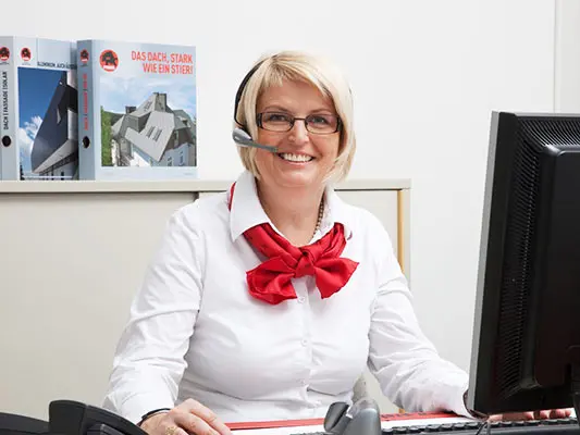 PREFA Mitarbeiterin mit weißer Bluse, rotem Schal und Headset am Schreibtisch.