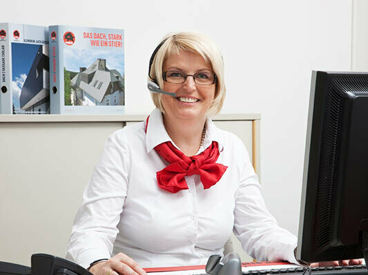 PREFA Mitarbeiterin mit weißer Bluse, rotem Schal und Headset am Schreibtisch.