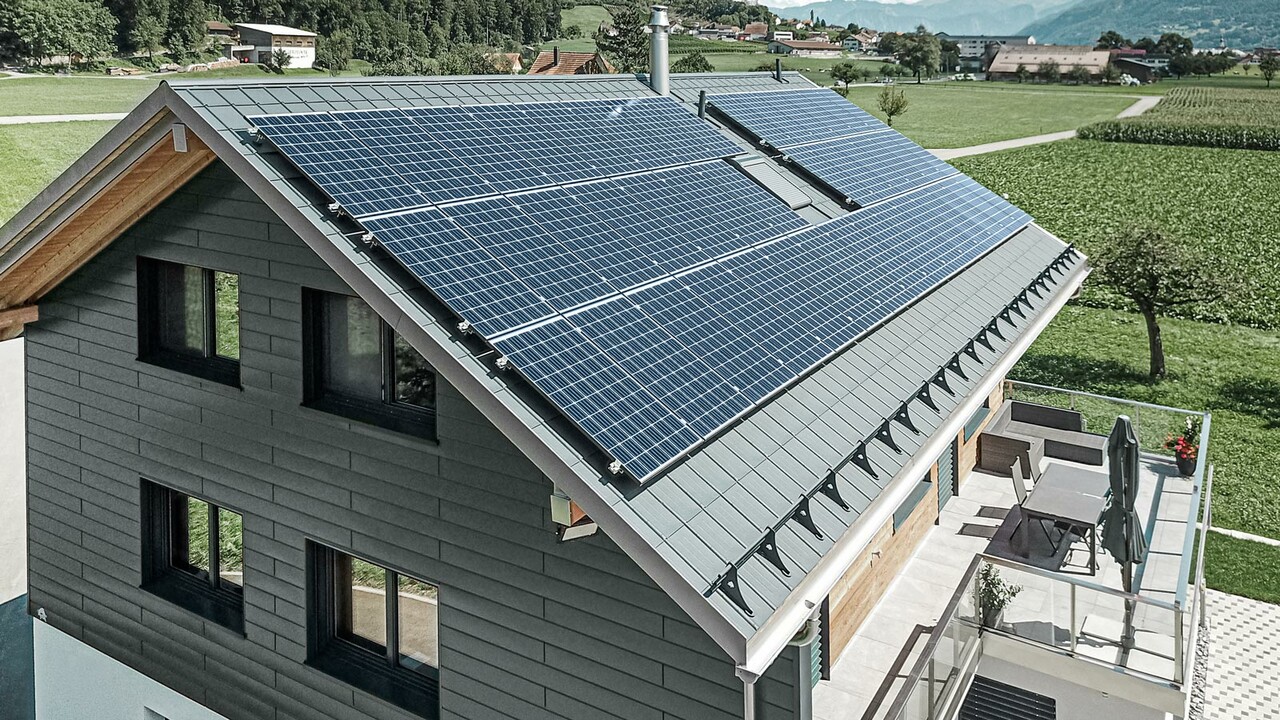 Das Bild zeigt ein modernes Einfamilienhaus mit mehreren PREFA Aluminiumprodukten. Die große Photovoltaikanlage (PV-Anlage) auf dem Dach verdeutlicht den Wert von nachhaltiger Stromerzeugung. Die PV-Module sind sorgfältig auf der gesamten Dachfläche installiert, um maximale Sonnenenergie zu nutzen. Zusätzlich ist ein Schneeschutzsystem integriert, das speziell für die Kombination mit Energiegewinnungsflächen entwickelt wurde. Dieses System verhindert das Abrutschen von Schnee und sorgt so für Schutz vor Dachlawinen. Auf dem Dach des Gebäudes wurde die bewährte PREFA Dachplatte in der Farbe Anthrazit verlegt. Das robuste Dachsystem aus Aluminium ist besonders langlebig und robust. Der Giebel des Hauses wurde mit hochwertigen PREFA Sidings in der gleichen Farbe verkleidet. Die Kombination der hochwertigen Blechverkleidungen bietet starken Schutz und eine eindrucksvolle Optik.