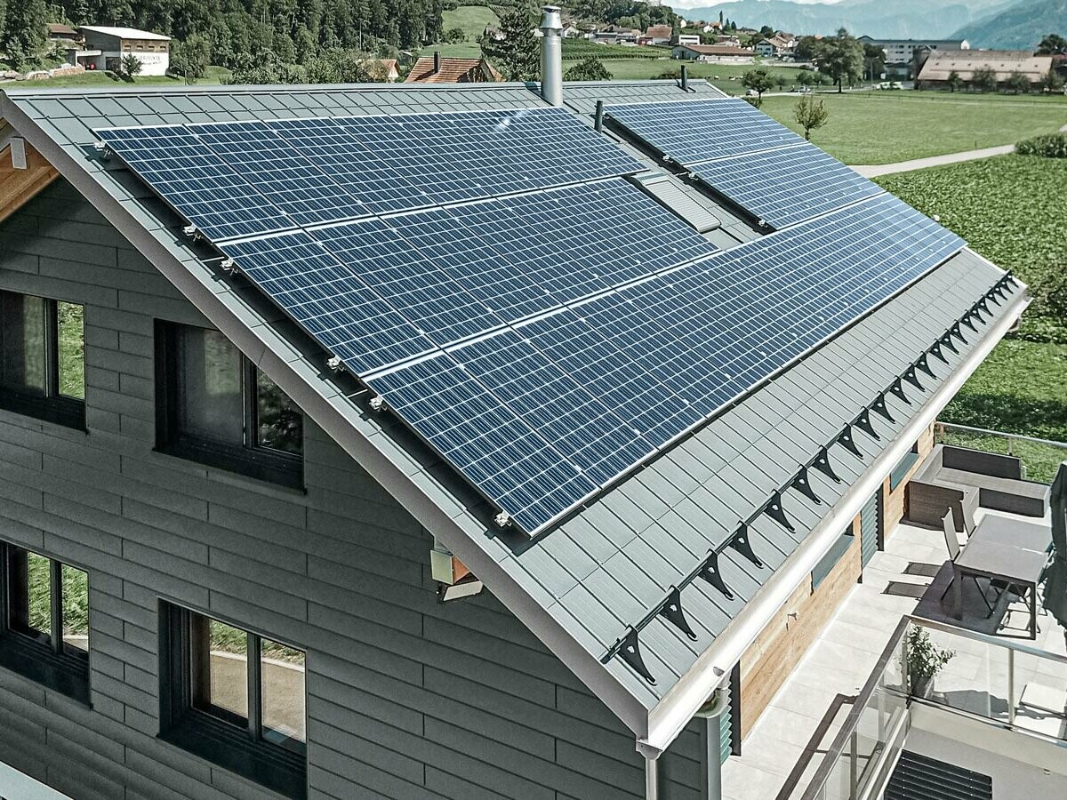 Das Bild zeigt ein modernes Einfamilienhaus mit mehreren PREFA Aluminiumprodukten. Die große Photovoltaikanlage (PV-Anlage) auf dem Dach verdeutlicht den Wert von nachhaltiger Stromerzeugung. Die PV-Module sind sorgfältig auf der gesamten Dachfläche installiert, um maximale Sonnenenergie zu nutzen. Zusätzlich ist ein Schneeschutzsystem integriert, das speziell für die Kombination mit Energiegewinnungsflächen entwickelt wurde. Dieses System verhindert das Abrutschen von Schnee und sorgt so für Schutz vor Dachlawinen. Auf dem Dach des Gebäudes wurde die bewährte PREFA Dachplatte in der Farbe Anthrazit verlegt. Das robuste Dachsystem aus Aluminium ist besonders langlebig und robust. Der Giebel des Hauses wurde mit hochwertigen PREFA Sidings in der gleichen Farbe verkleidet. Die Kombination der hochwertigen Blechverkleidungen bietet starken Schutz und eine eindrucksvolle Optik.