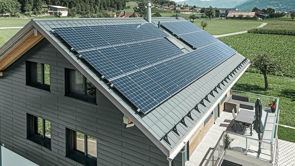 Das Bild zeigt ein modernes Einfamilienhaus mit mehreren PREFA Aluminiumprodukten. Die große Photovoltaikanlage (PV-Anlage) auf dem Dach verdeutlicht den Wert von nachhaltiger Stromerzeugung. Die PV-Module sind sorgfältig auf der gesamten Dachfläche installiert, um maximale Sonnenenergie zu nutzen. Zusätzlich ist ein Schneeschutzsystem integriert, das speziell für die Kombination mit Energiegewinnungsflächen entwickelt wurde. Dieses System verhindert das Abrutschen von Schnee und sorgt so für Schutz vor Dachlawinen. Auf dem Dach des Gebäudes wurde die bewährte PREFA Dachplatte in der Farbe Anthrazit verlegt. Das robuste Dachsystem aus Aluminium ist besonders langlebig und robust. Der Giebel des Hauses wurde mit hochwertigen PREFA Sidings in der gleichen Farbe verkleidet. Die Kombination der hochwertigen Blechverkleidungen bietet starken Schutz und eine eindrucksvolle Optik.