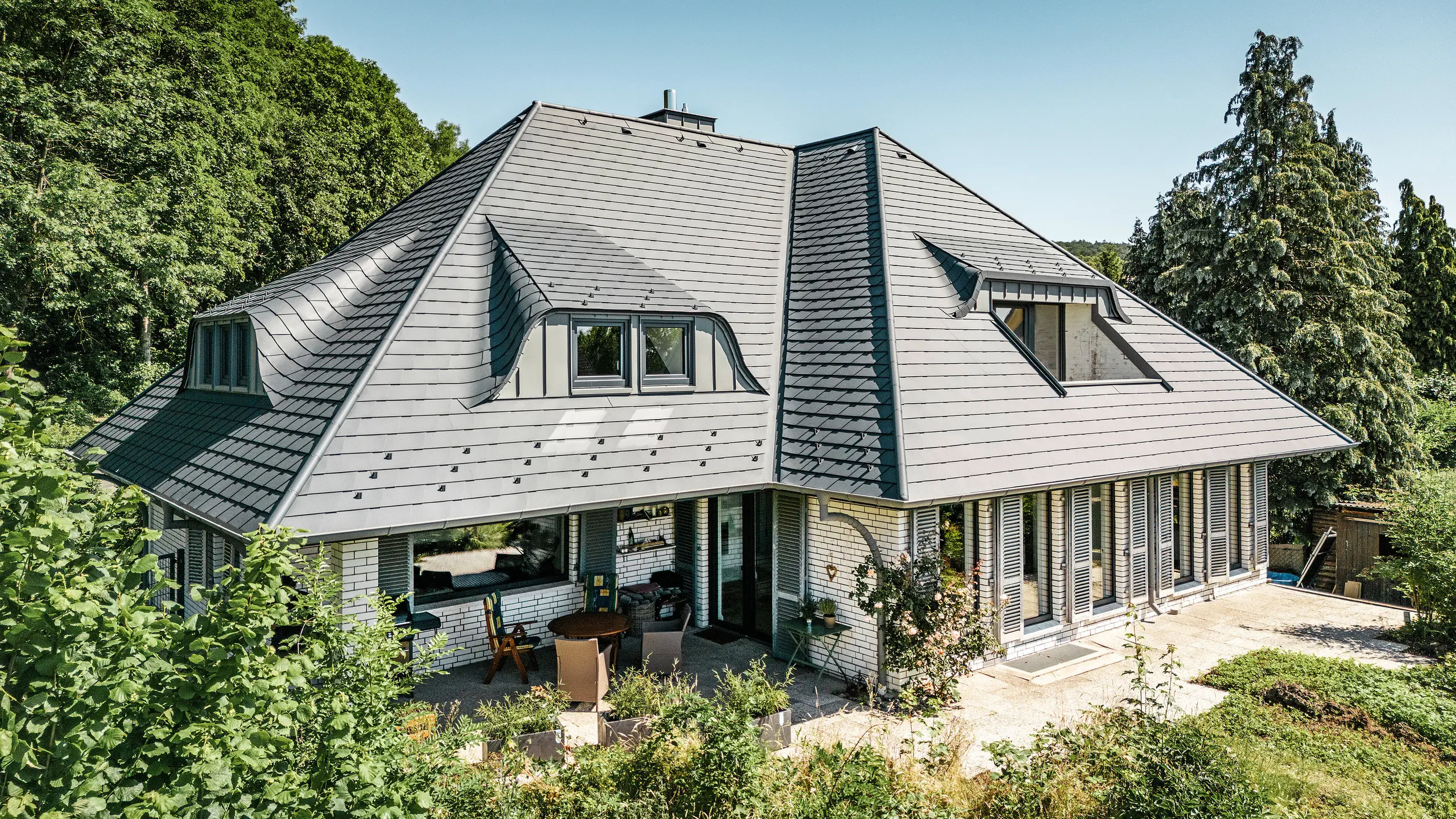 Modernes Einfamilienhaus in Einbeck, Deutschland, mit PREFA Dachschindeln in P.10 Anthrazit. Die langlebigen PREFA Dachschindeln aus Aluminium sind für ihre Qualität und Langlebigkeit bekannt. Die Dachgestaltung des Gebäudes betont die individuelle Architektur mit Hechtgauben, die durch die Flexibilität der PREFA Dachschindeln hervorgehoben wird. Die Dachentwässerung in Form des PREFA Ablaufrohrs bzw. der Hängerinne ist ebenfalls in Anthrazit gehalten. Rund um das Einfamilienhaus ist ein großzügiger und gepflegter Garten sowie eine Terrasse zu sehen. Hinter dem Haus liegt ein Wald, der - gemeinsam mit dem blauen Himmel - eine natürliche Atmosphäre schafft. 