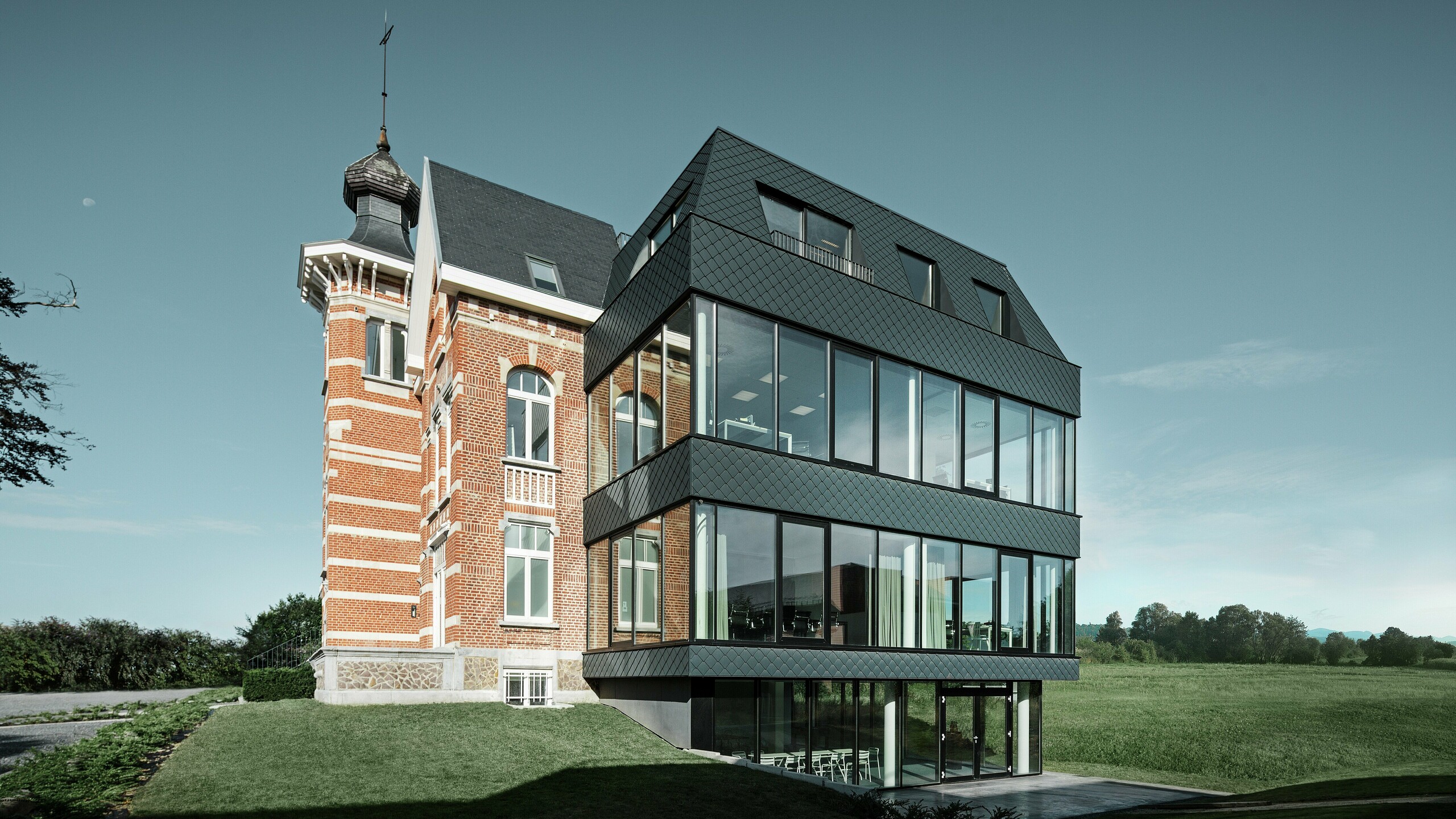 Seitlicher Blick auf das Kasteeltje Heizel, wo historischer Backsteinbau mit Turm nahtlos in einen modernen Anbau übergeht. Die anthrazitfarbenen PREFA Wandrauten setzen mit ihrer markanten Struktur elegante Kontraste zu den großzügigen Glasflächen. Die klare Linienführung des Neubaus verstärkt die Verbindung von Tradition und moderner Architektur in einer eindrucksvollen Kulisse.