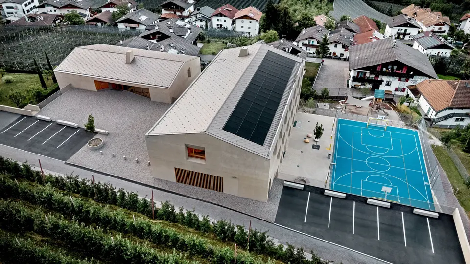 Luftaufnahme des Bildungszentrums Riffian in Südtirol mit Kindergarten, Volksschule und Sportfläche. Die PREFA Dachrauten 44 × 44 in P.10 Bronze bilden eine präzise, langlebige Aluminiumdeckung und fügen sich harmonisch in die alpine Umgebung ein. Die moderne Architektur zeigt wetterfesten Schutz und klare Gestaltung im Detail.