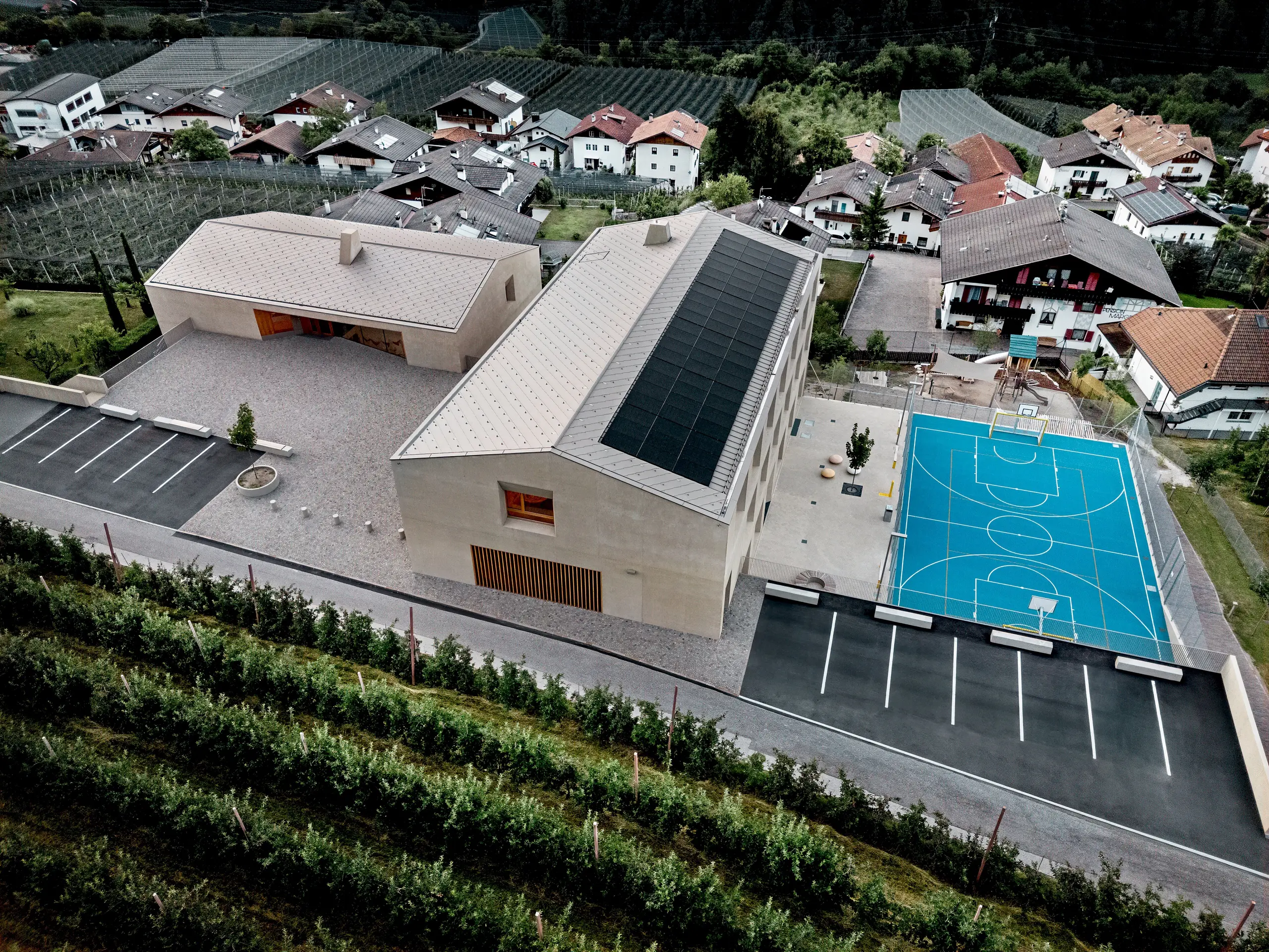 Luftaufnahme des Bildungszentrums Riffian in Südtirol mit Kindergarten, Volksschule und Sportfläche. Die PREFA Dachrauten 44 × 44 in P.10 Bronze bilden eine präzise, langlebige Aluminiumdeckung und fügen sich harmonisch in die alpine Umgebung ein. Die moderne Architektur zeigt wetterfesten Schutz und klare Gestaltung im Detail.