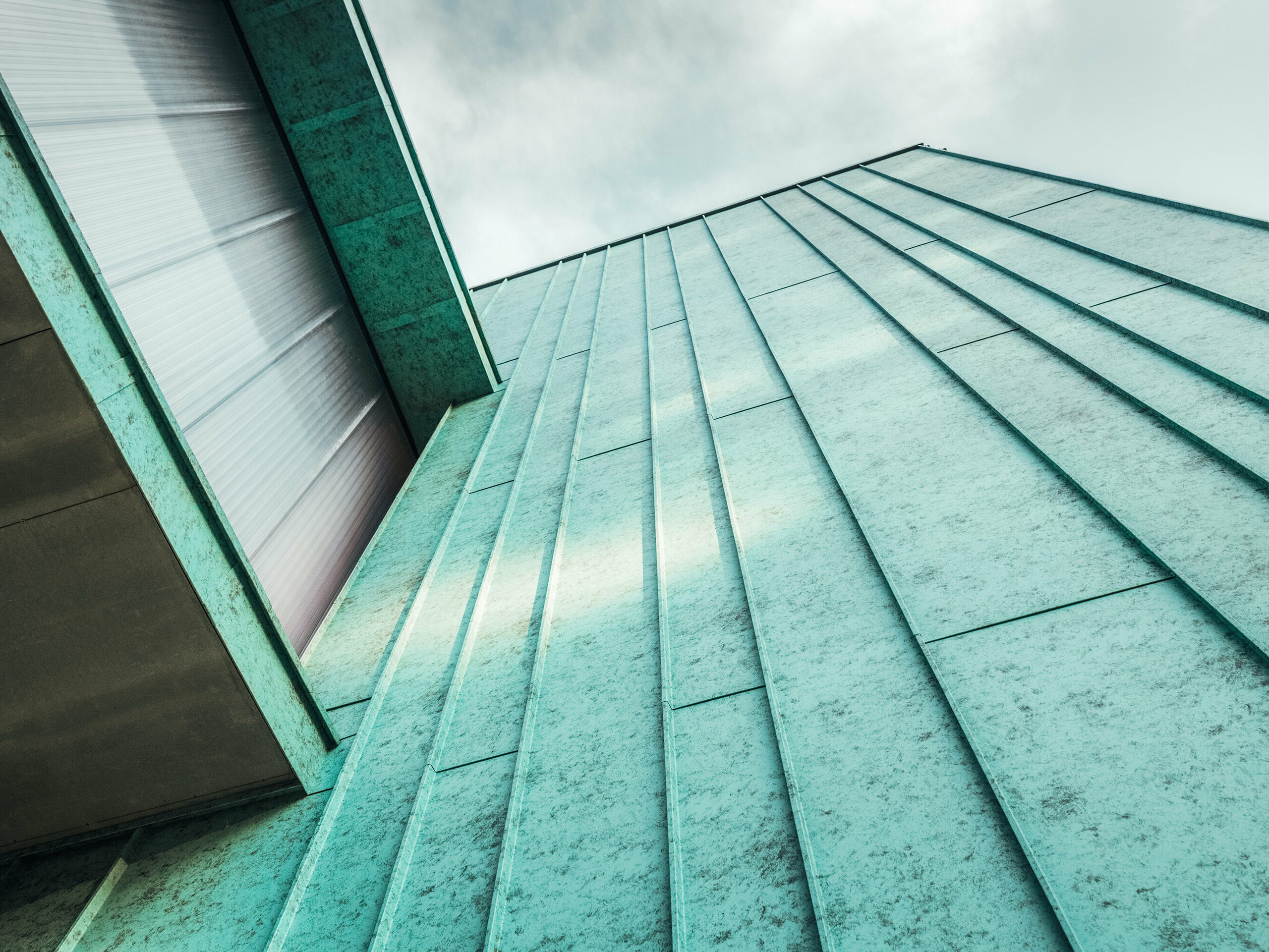 Nahaufnahme einer modernen Fassade, die mit PREFALZ in P.10 Patinagrün verkleidet ist. Die Aufnahme aus einer Perspektive von unten nach oben betont die klaren Linien der vertikalen Stehfalzdeckung und die einheitliche Farbgebung. Die Textur der Fassade verleiht dem Gebäude Charakter, während das reflektierte Licht und die Schatten den metallischen Glanz des Aluminiums hervorheben. Die besondere Farbbeschichtung fügt sich harmonisch in die Umgebung ein und schafft ein modernes, ästhetisch ansprechendes Erscheinungsbild.