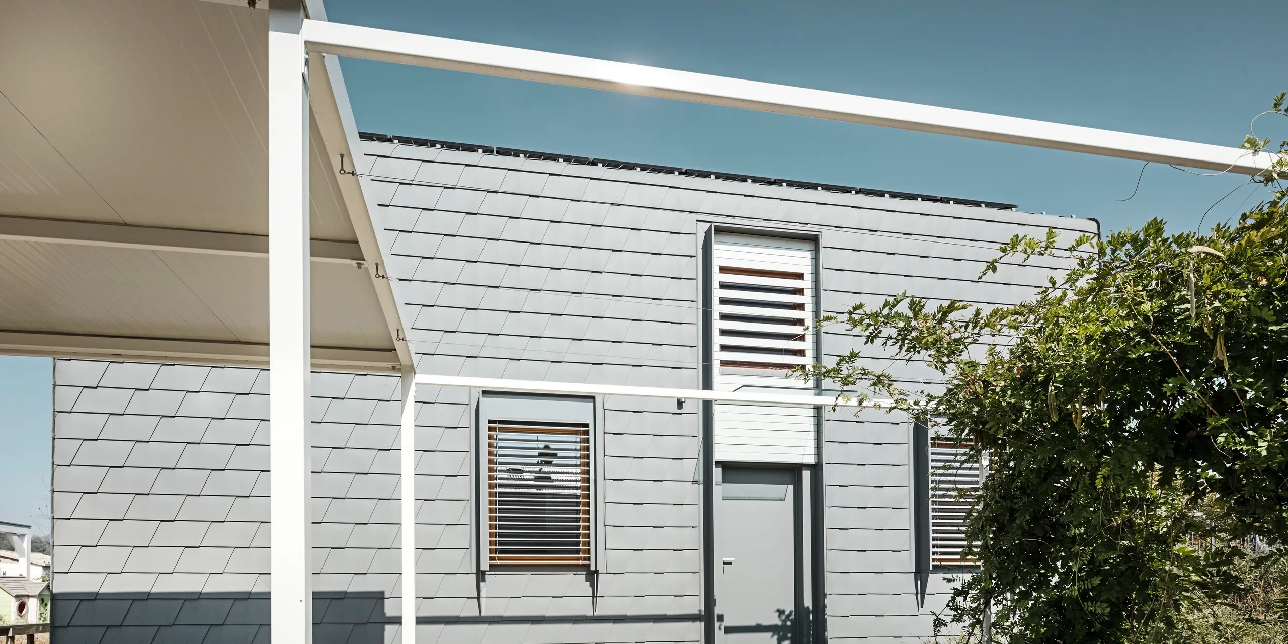 Seitenansicht eines modernen Einfamilienhauses, dessen Dach und Fassade mit PREFA Dach- und Wandschindeln in Hellgrau verkleidet sind. Die schindelbedeckten Flächen zeigen ein sauberes, geometrisches Muster, das für eine strukturierte und zeitgenössische Ästhetik sorgt. Die Schindeln komplementieren die klaren weißen Rahmen und Akzente des Gebäudes, während im Vordergrund ein Vordach und üppiges Grün für einen angenehmen Kontrast sorgen.