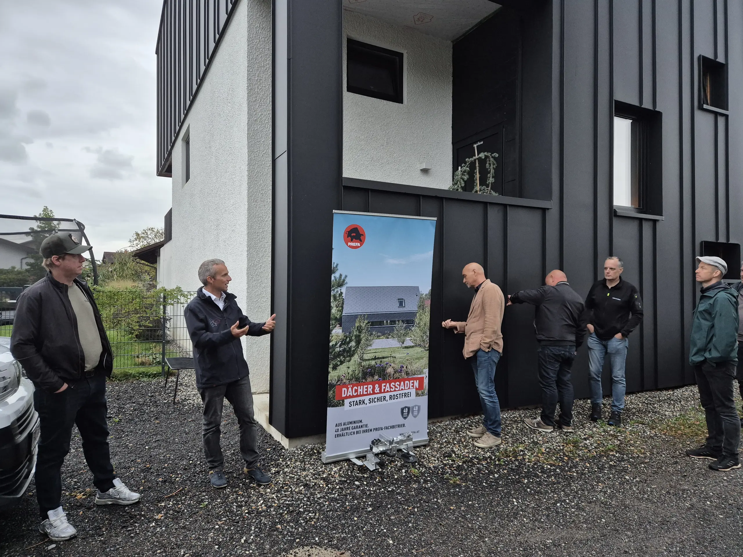 Vor einer markanten, dunkel verkleideten Fassade erläutert Markus Metzler, PREFA Objektberater, den Teilnehmenden der PREFA Objekttour die architektonischen Besonderheiten des sanierten Wohnhauses. Eyecatcher ist die elegante PREFA Aluminiumfassade mit ihrer klaren Linienführung und modernen Optik. Einige Gäste prüfen die Oberfläche aus nächster Nähe und erleben die hochwertige Verarbeitung sowie die langlebige Qualität der PREFA Lösungen hautnah.