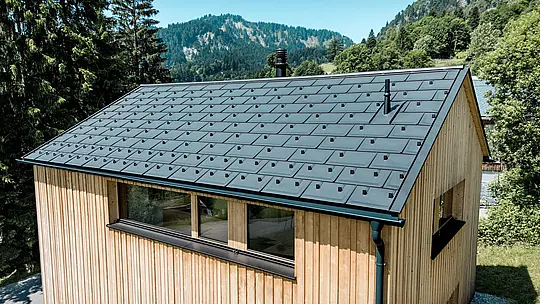 Im hellen Bergsommer rückt das schwarze PREFA Aluminiumdach von Haus Immi eindrucksvoll in den Mittelpunkt. Die flächenbündig integrierten Solardachplatten bilden mit den Dachplatten R.16 in P.10 Schwarz eine ruhige, homogene Dachhaut. Das leichte, robuste Aluminiumsystem erzeugt Solarstrom und schützt das Holzhaus zuverlässig vor anspruchsvollem alpinem Wetter sowie hohen Schneelasten.