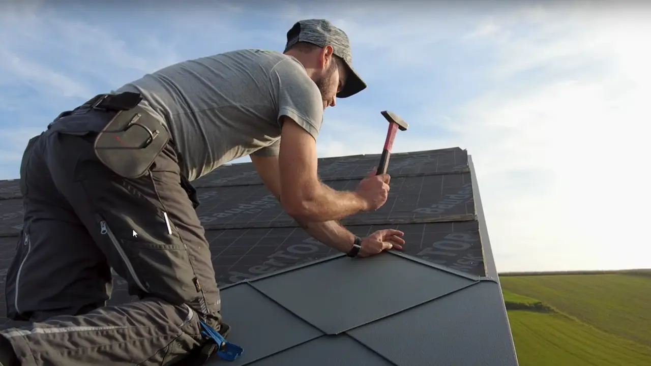 Spengler in Aktion: Mit einem Hammer verlegt der Eigentümer der Firma „Der Blechmann“ präzise die PREFA Dachraute 44 × 44. Die Präzision bei der Installation ist bei den bereits verlegten Rauten am unteren Bildrand zu erkennen. Der Mann trägt eine Mütze und kniet mit dem Rücken zur Kamera auf dem Dach. Im Hintergrund ist der klare Himmel und eine grüne Landschaft zu erkennen. Der harmonische Hintergrund unterstreicht die Symbolik des Bildes: Nachhaltiges und langlebiges Handwerk mit hochwertigen PREFA Aluminiumprodukten.