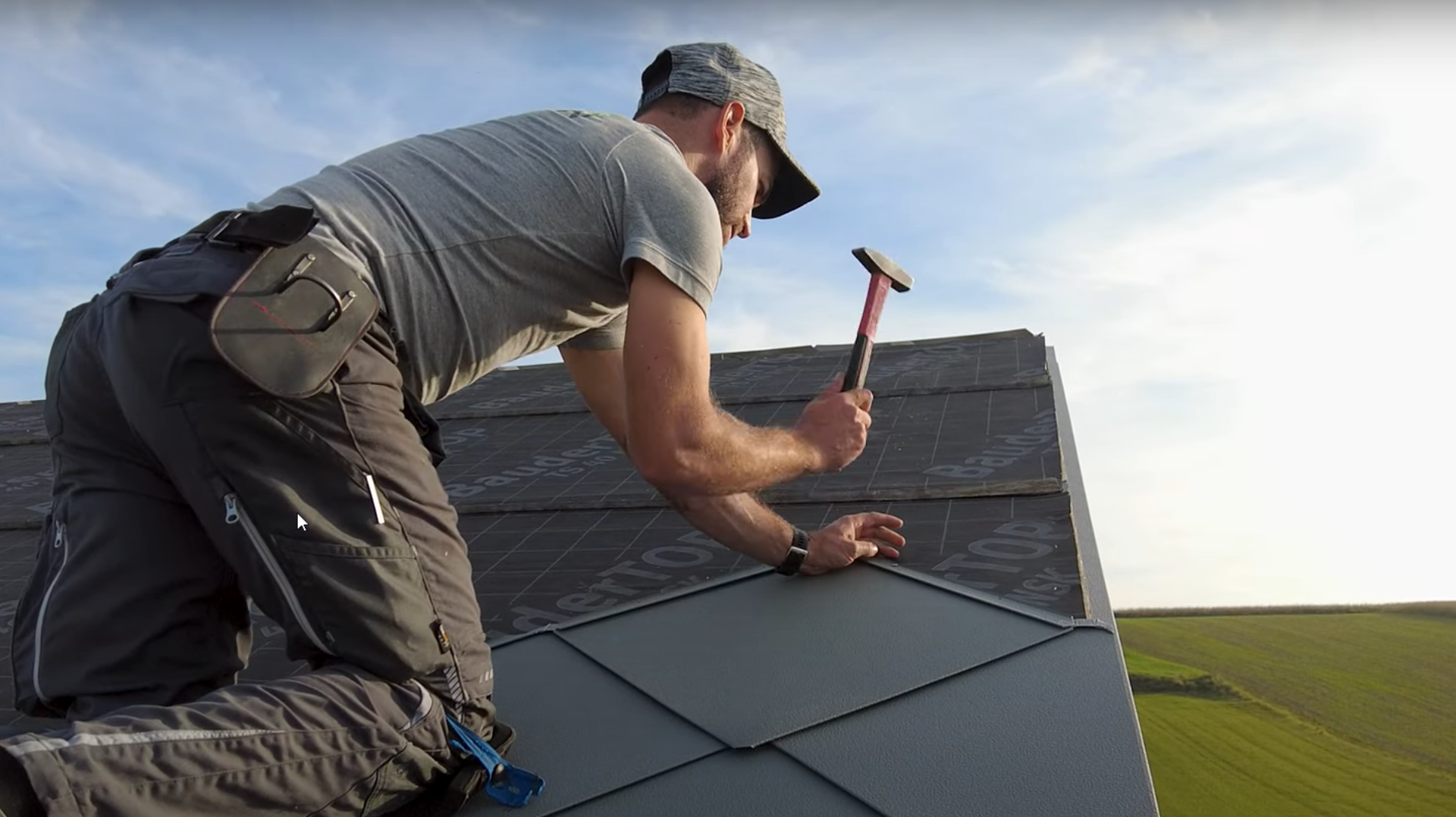 Spengler in Aktion: Mit einem Hammer verlegt der Eigentümer der Firma „Der Blechmann“ präzise die PREFA Dachraute 44 × 44. Die Präzision bei der Installation ist bei den bereits verlegten Rauten am unteren Bildrand zu erkennen. Der Mann trägt eine Mütze und kniet mit dem Rücken zur Kamera auf dem Dach. Im Hintergrund ist der klare Himmel und eine grüne Landschaft zu erkennen. Der harmonische Hintergrund unterstreicht die Symbolik des Bildes: Nachhaltiges und langlebiges Handwerk mit hochwertigen PREFA Aluminiumprodukten.